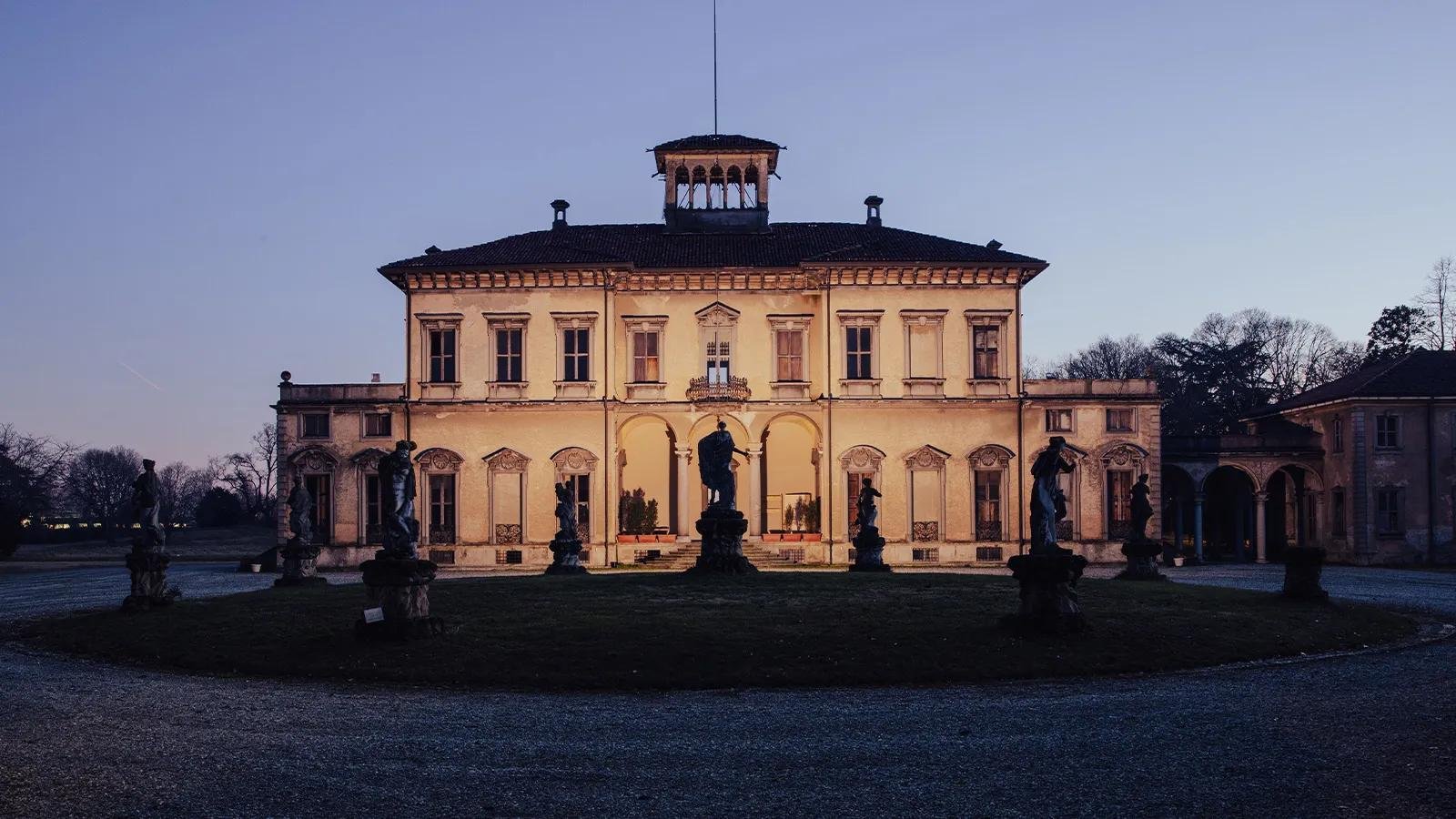 A villa lit at dusk, with statues standing along the garden in front of it.