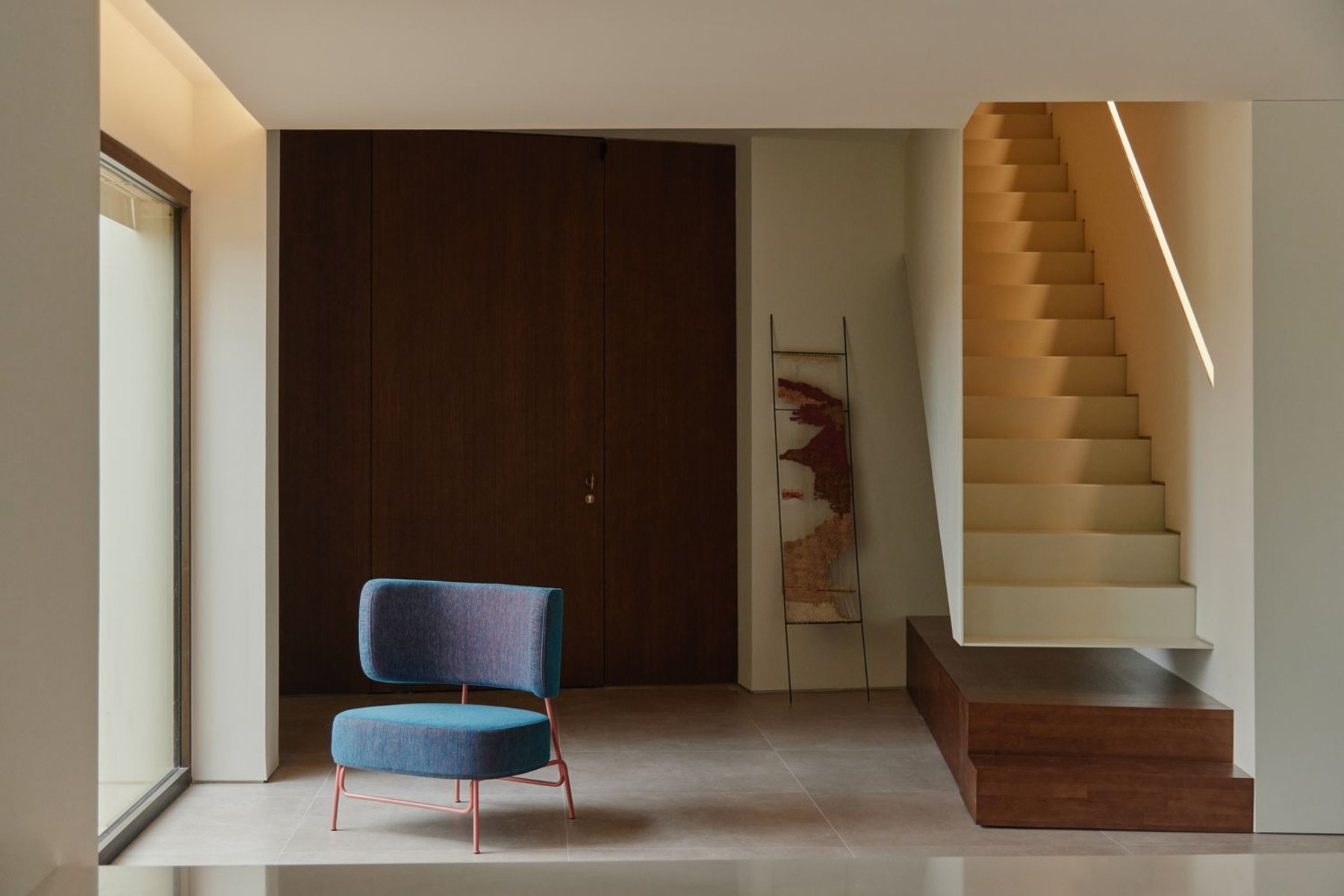 An entrance hall with a blue armchair, a dark wall and a wooden stair.