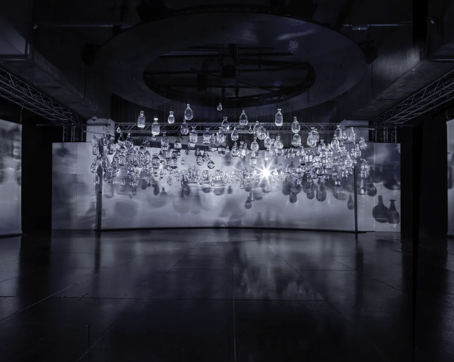 A wall of hanging glass lamps lit in a dark hall, reflected in the polished floor.