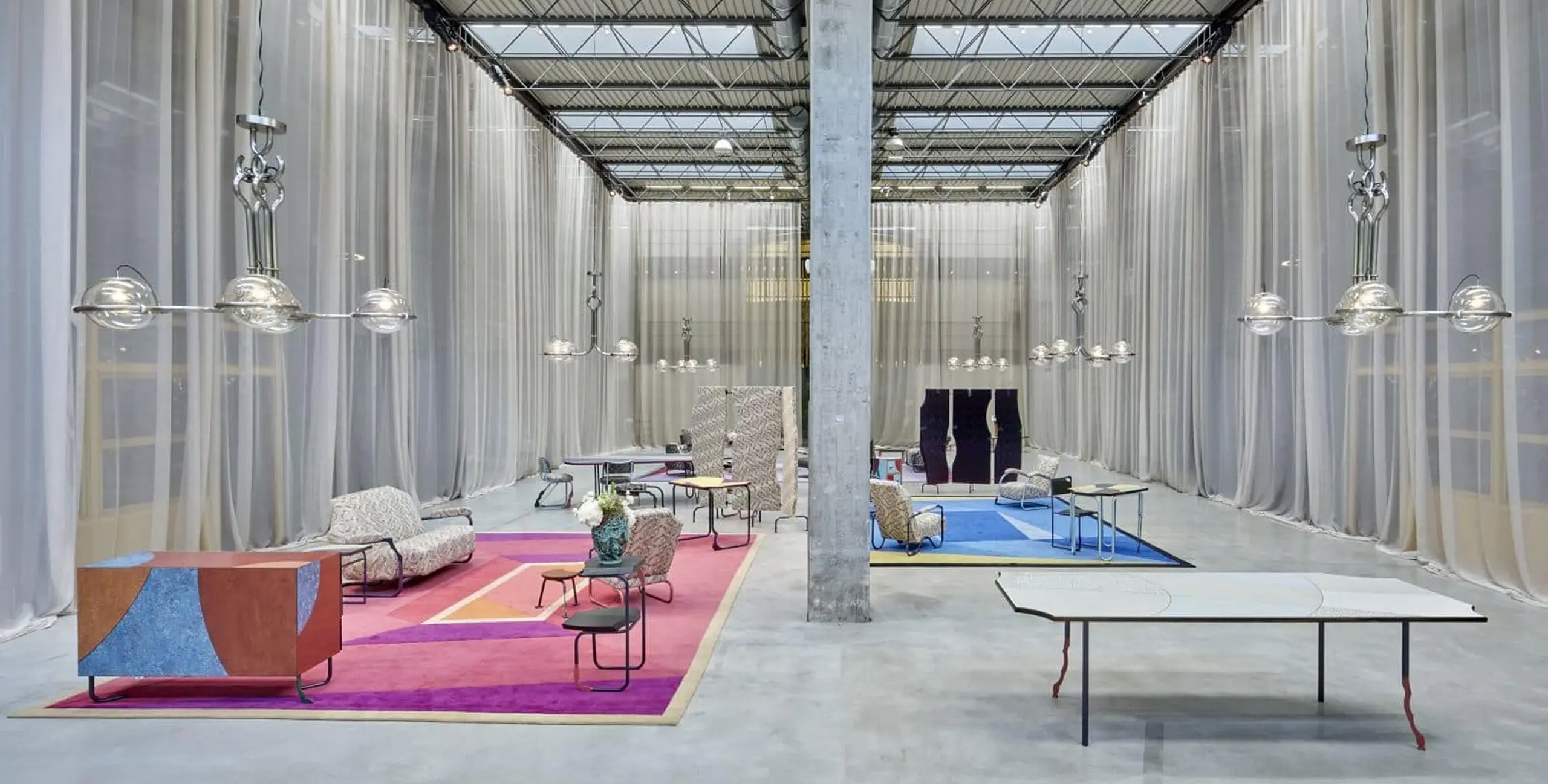 A design fair in a factory hall, with pink and blue carpets and sheer curtains between the columns.