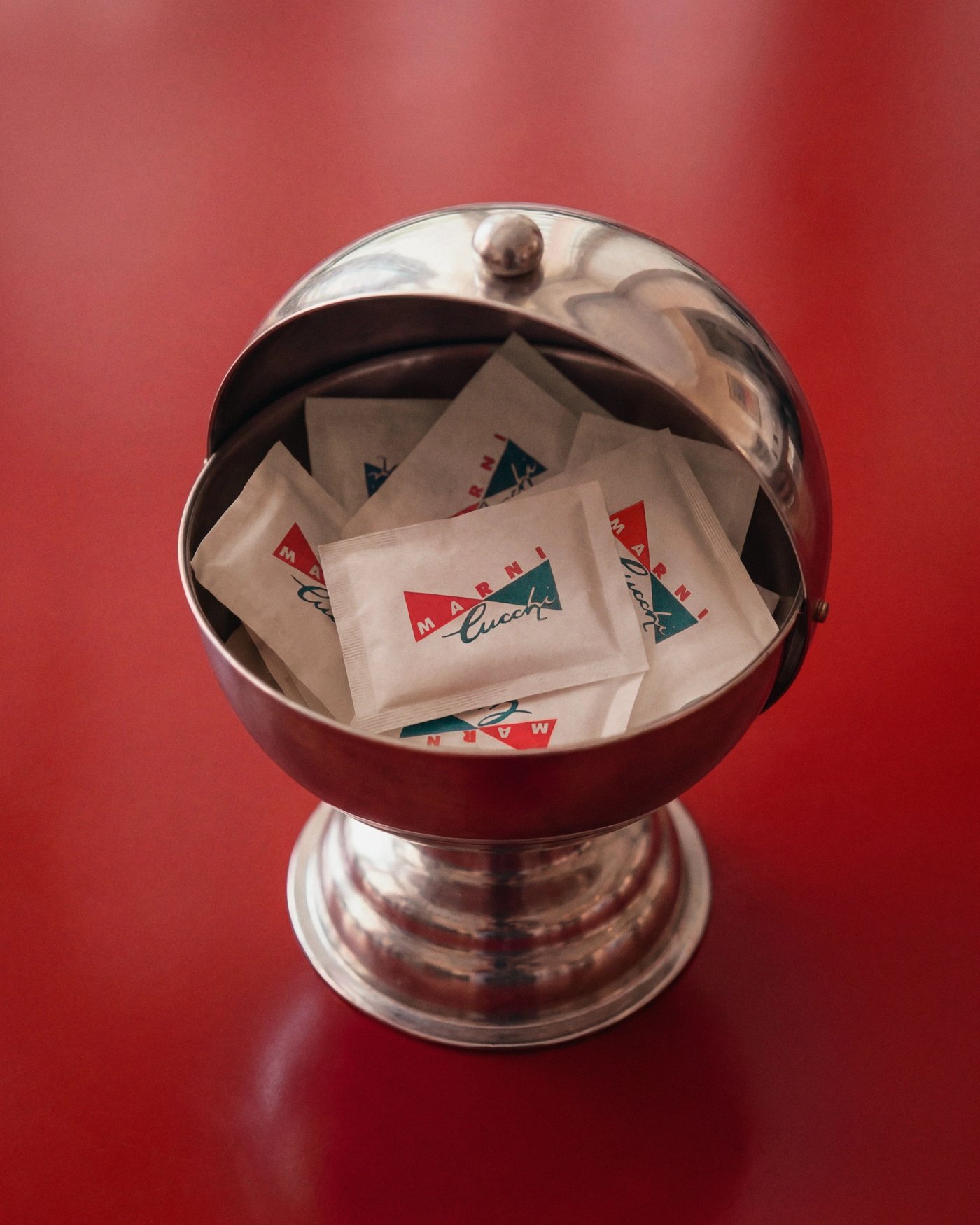 A chrome sugar bowl filled with Cucchi sachets, on a red table.