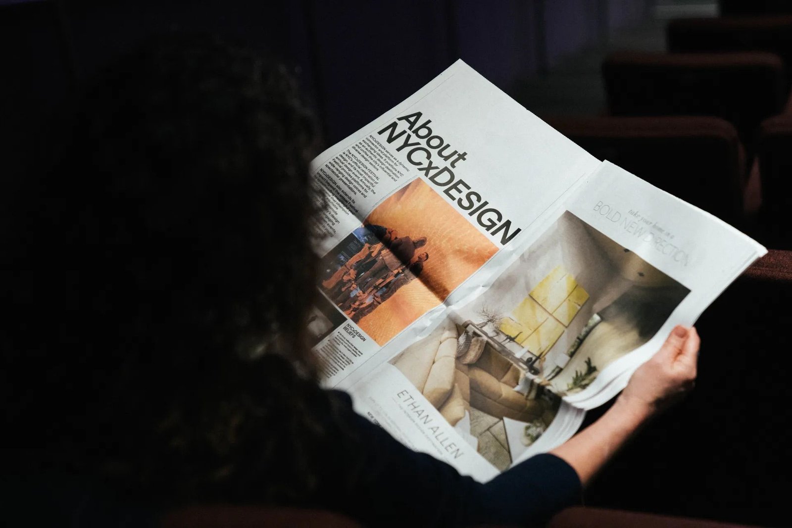 A woman reads the About NYCxDESIGN newspaper in a dark room.
