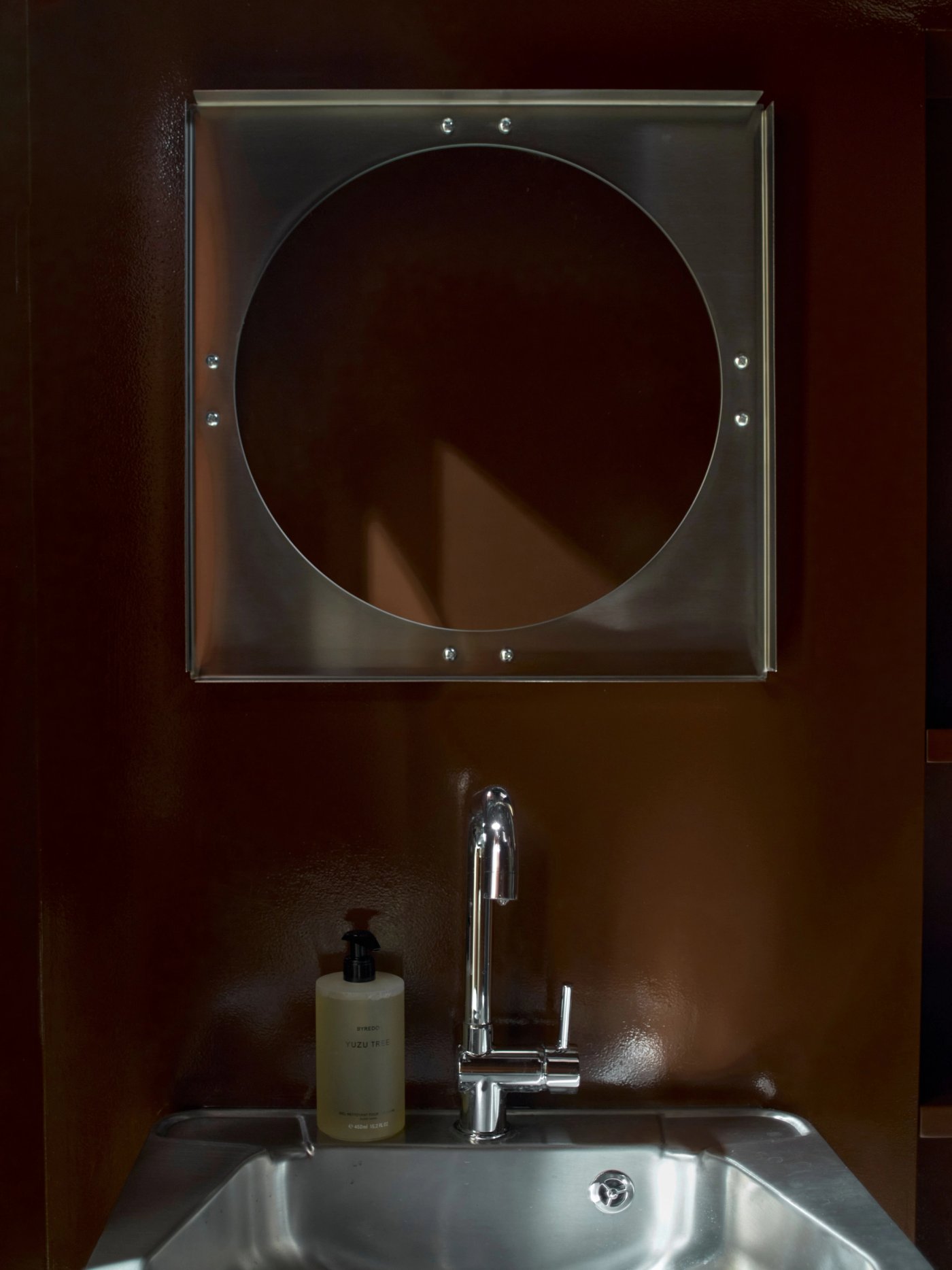 A round steel mirror above a steel sink, on a deep brown wall.