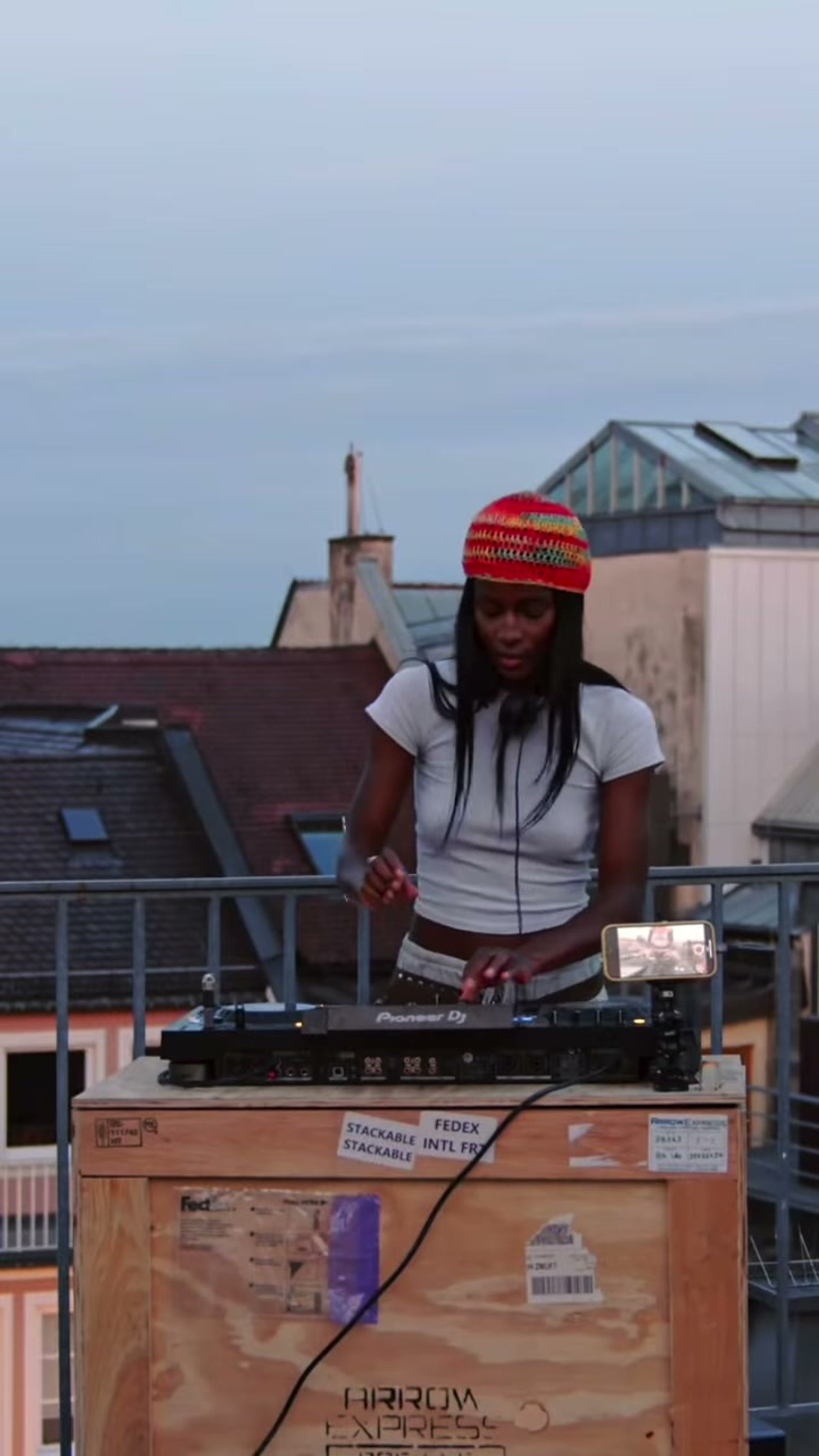 A DJ in a red knit beanie plays a set on a Munich rooftop at dusk.