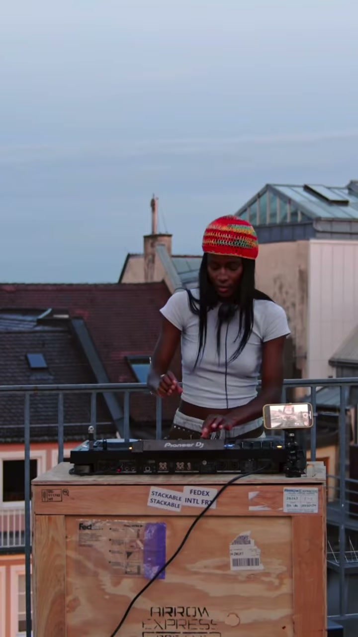 A DJ in a red knit beanie plays a set on a Munich rooftop at dusk.