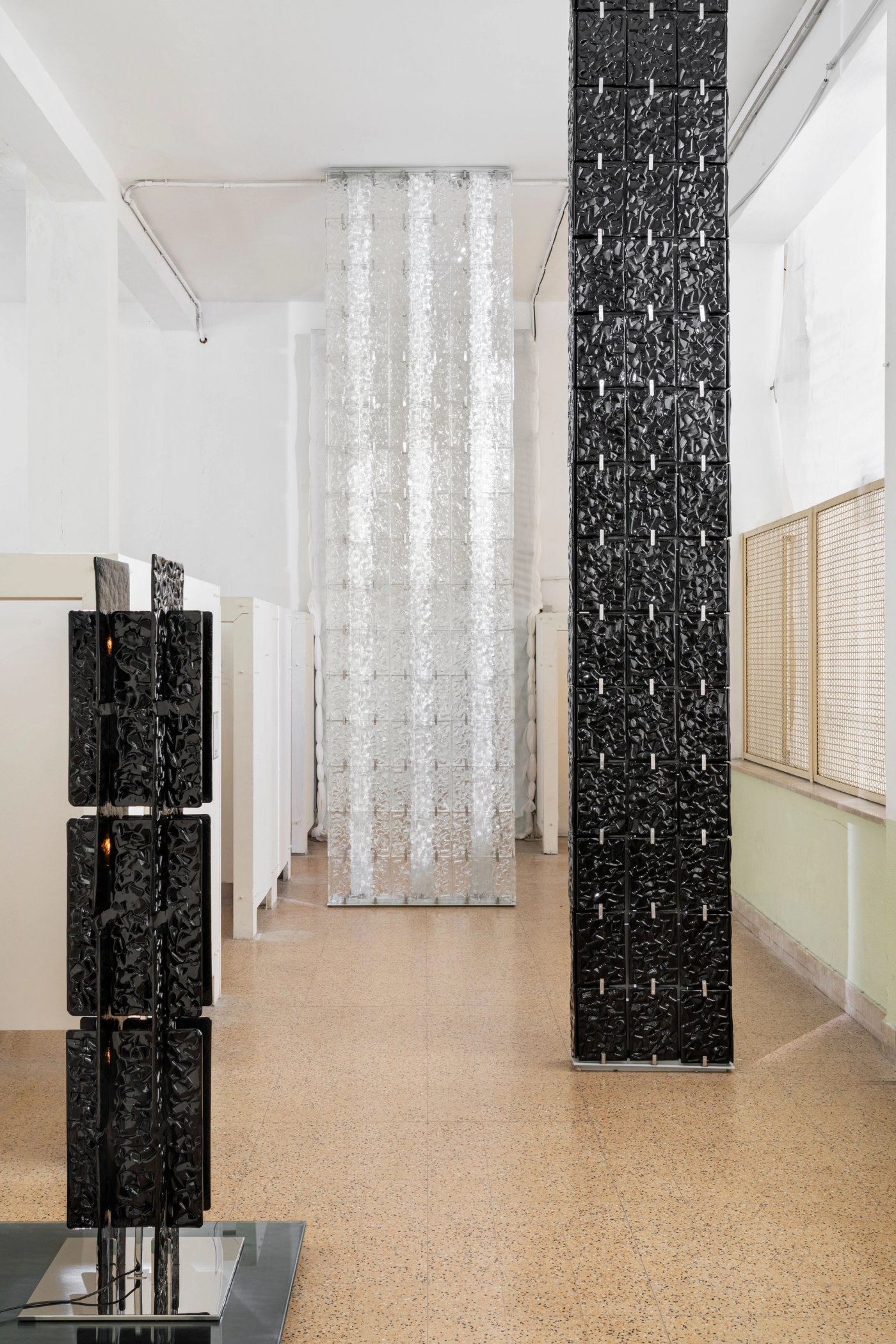 Columns of black cast glass bricks in a gallery, with a curtain of clear glass bricks behind them.