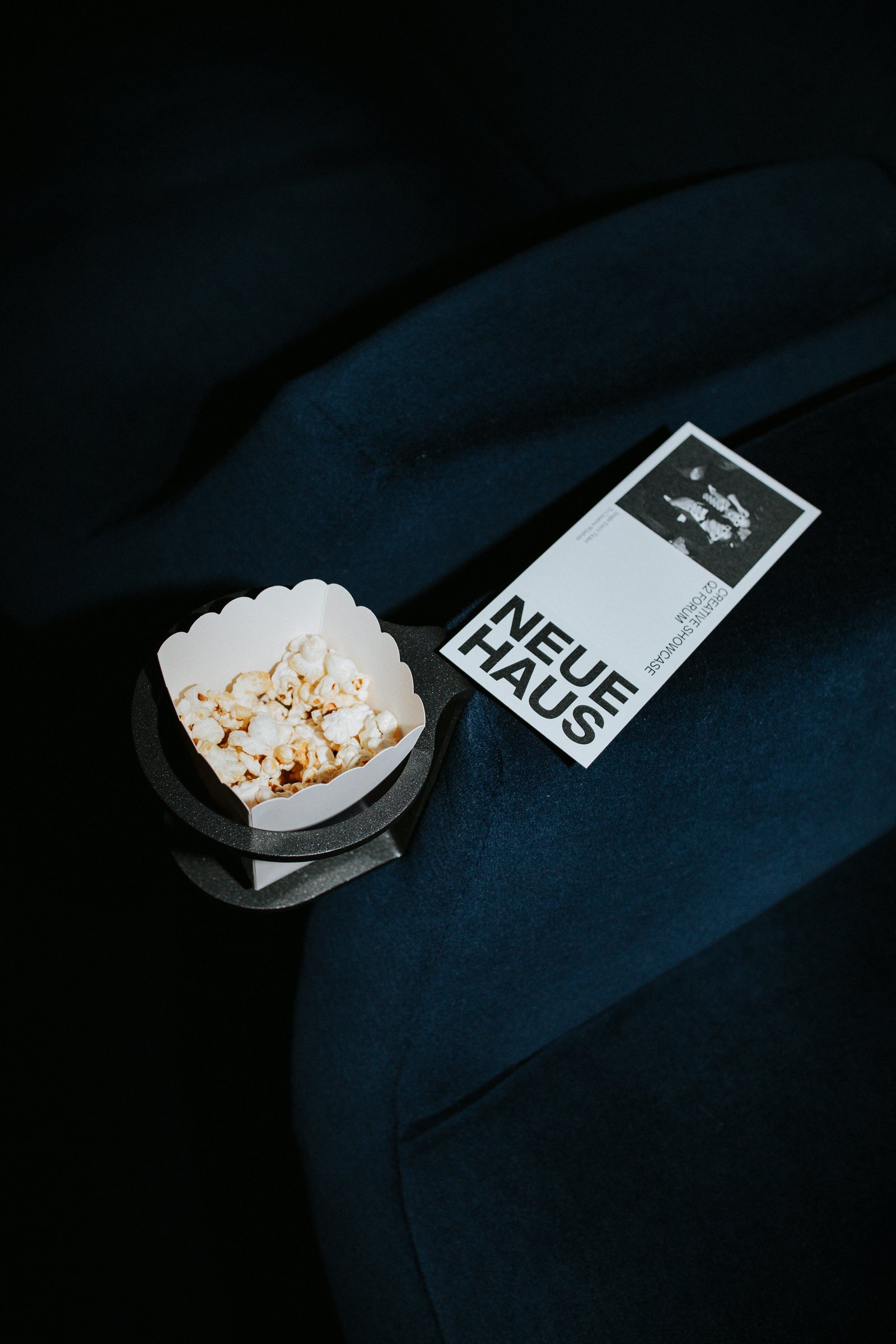 A NeueHaus card and a paper cup of popcorn on a dark blue velvet seat.