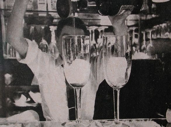 A black and white photograph of a barman pouring a drink into tall glasses.
