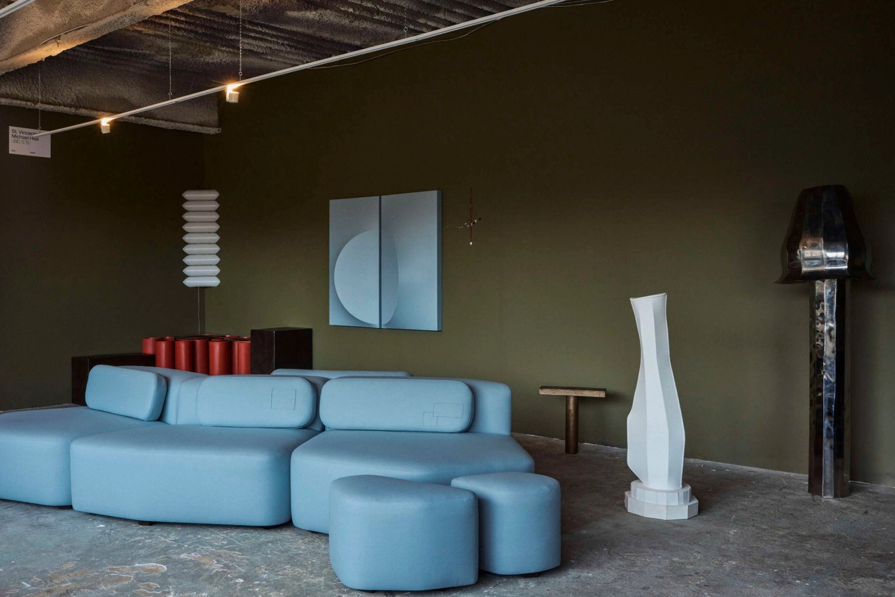A lounge with pale blue sofas against a dark green wall, under exposed lights.