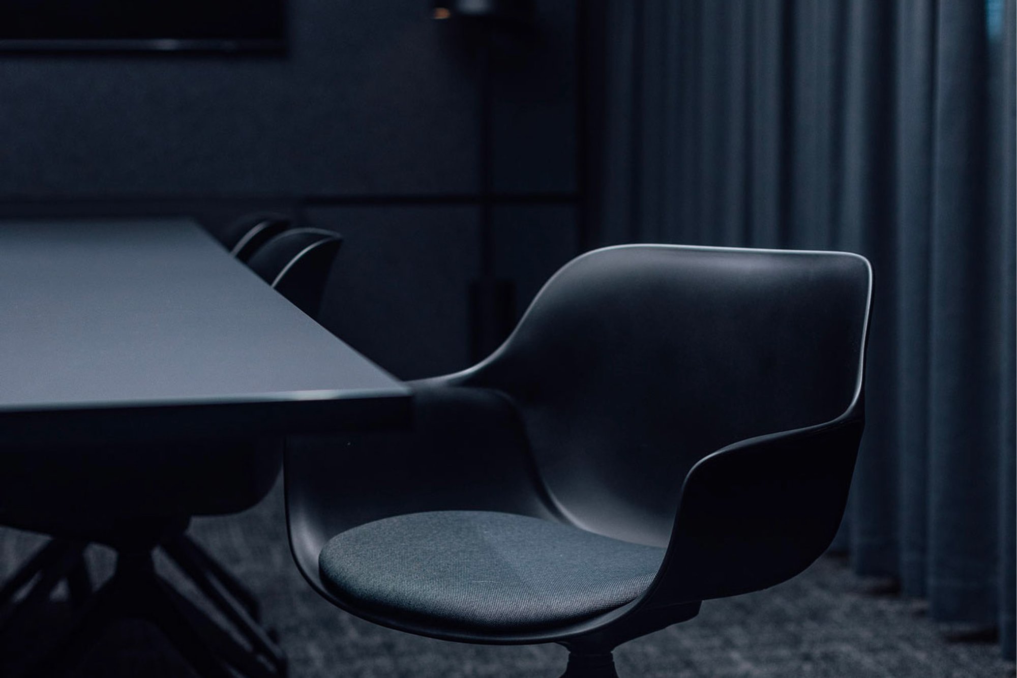 A dark meeting room with a moulded chair at the end of a long table.