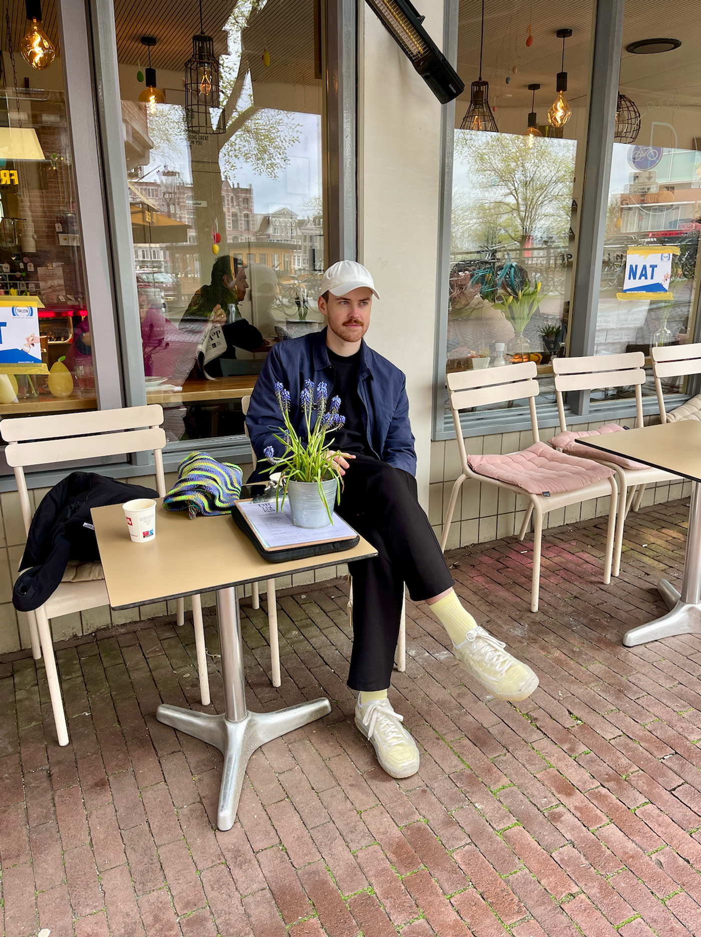 A man sits at a cafe table on an Amsterdam street, with a plant and a coffee in front of him.