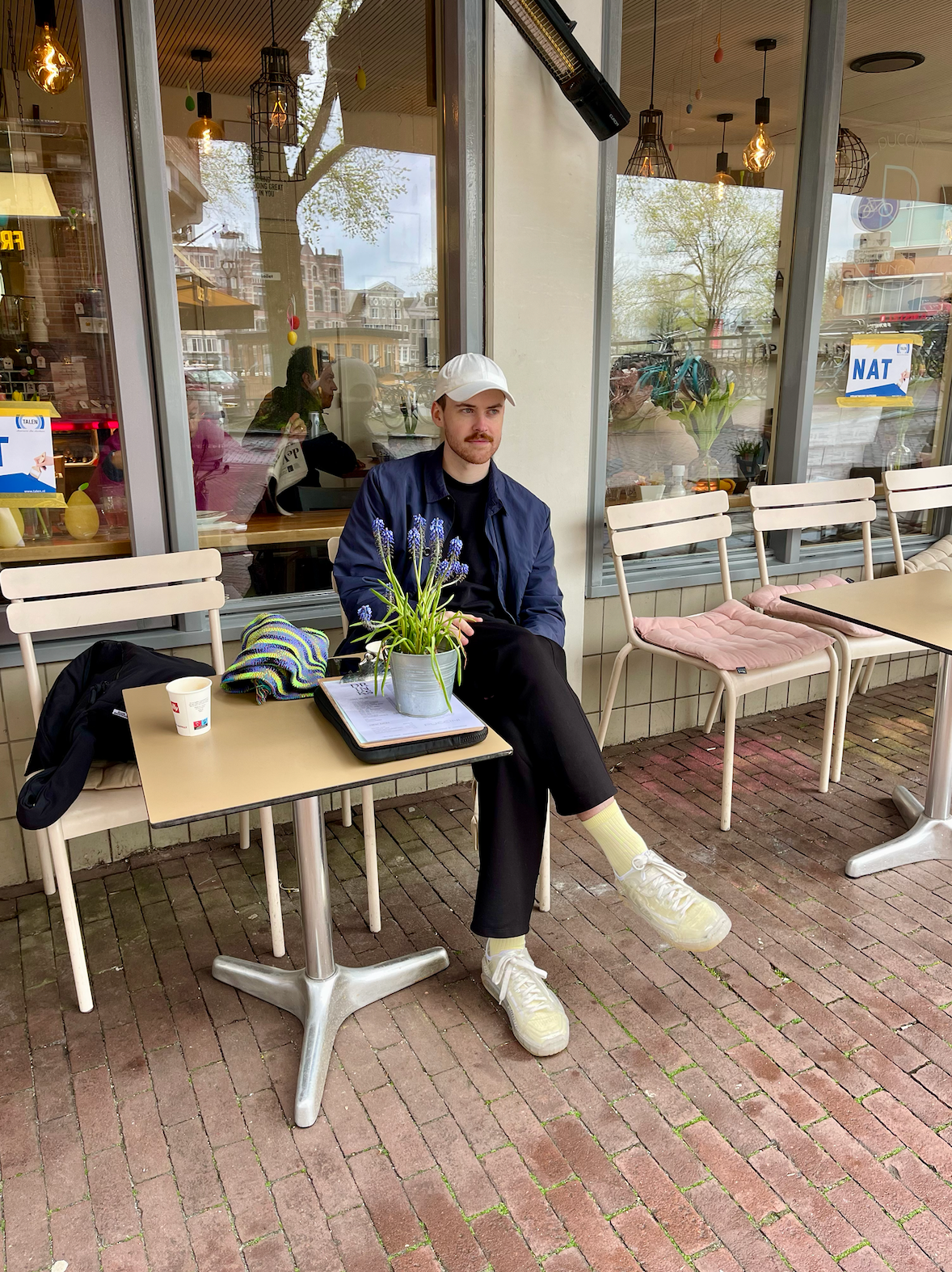 A man sits at a cafe table on an Amsterdam street, with a plant and a coffee in front of him.