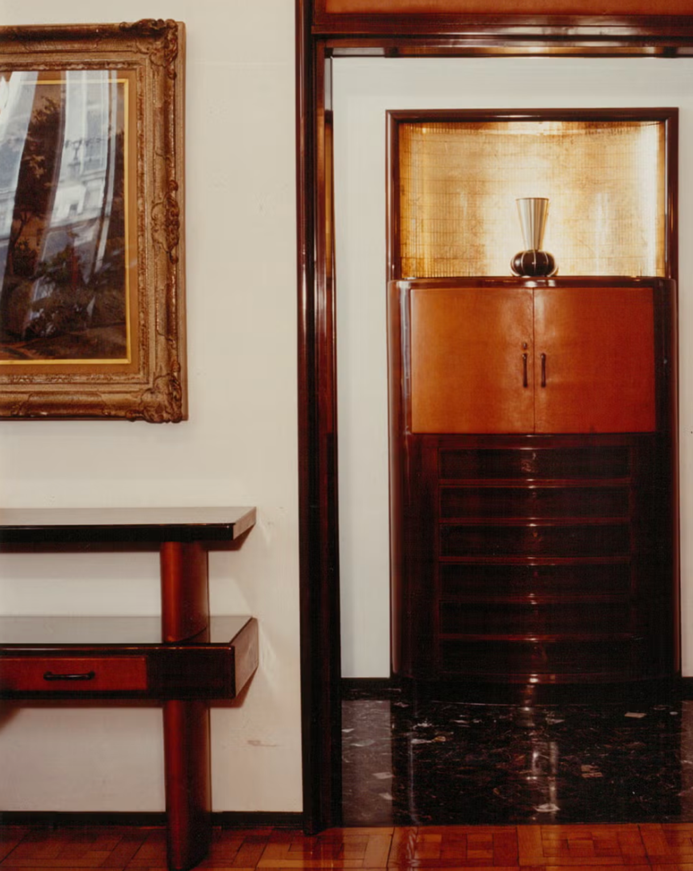 A polished wood cabinet and a console table in a Milanese apartment, beside a gilt framed landscape painting.
