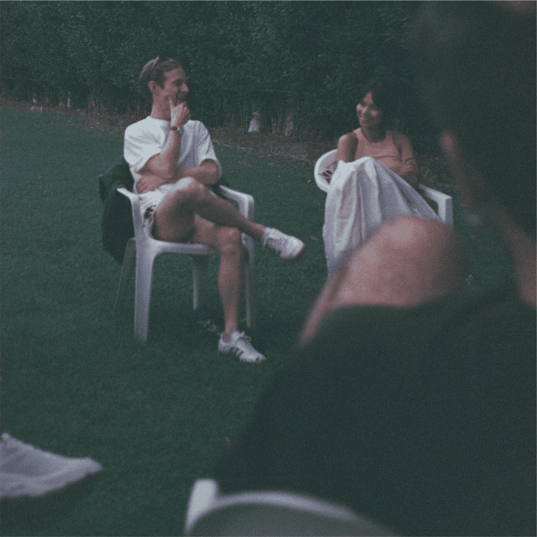 Two people talk in garden chairs on the grass at dusk, watched by the group around them.
