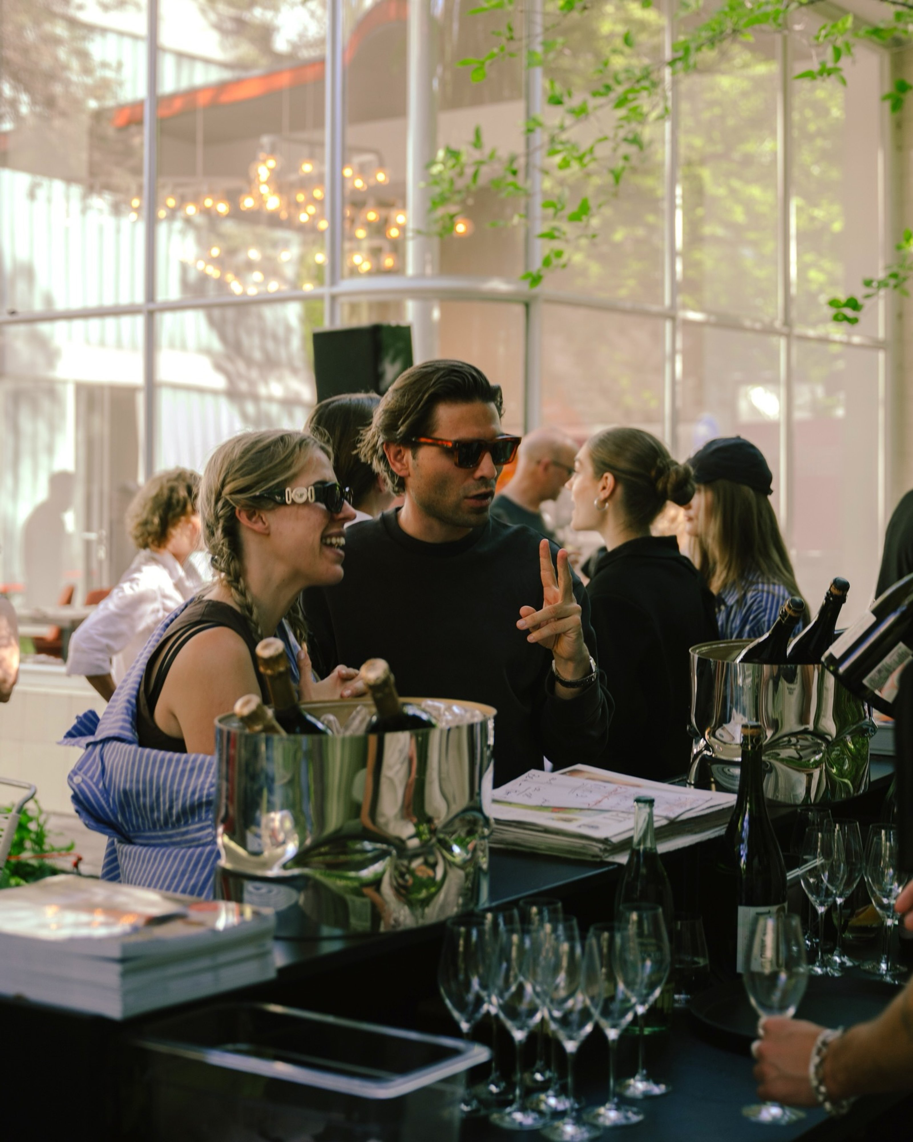 Guests in sunglasses talk at a bar lined with ice buckets of sparkling wine in a glass-walled gallery.