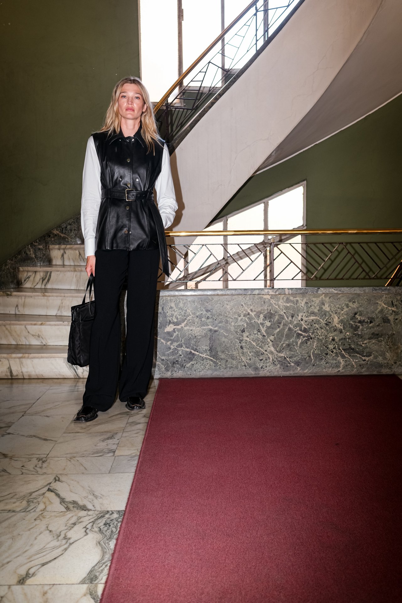 A woman stands in a marble foyer beside a staircase, on a red carpet runner.