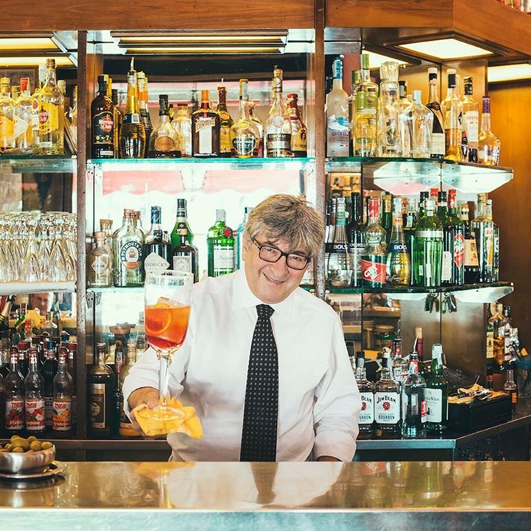 A barman in a white shirt and tie holds up a Negroni Sbagliato behind the bar.