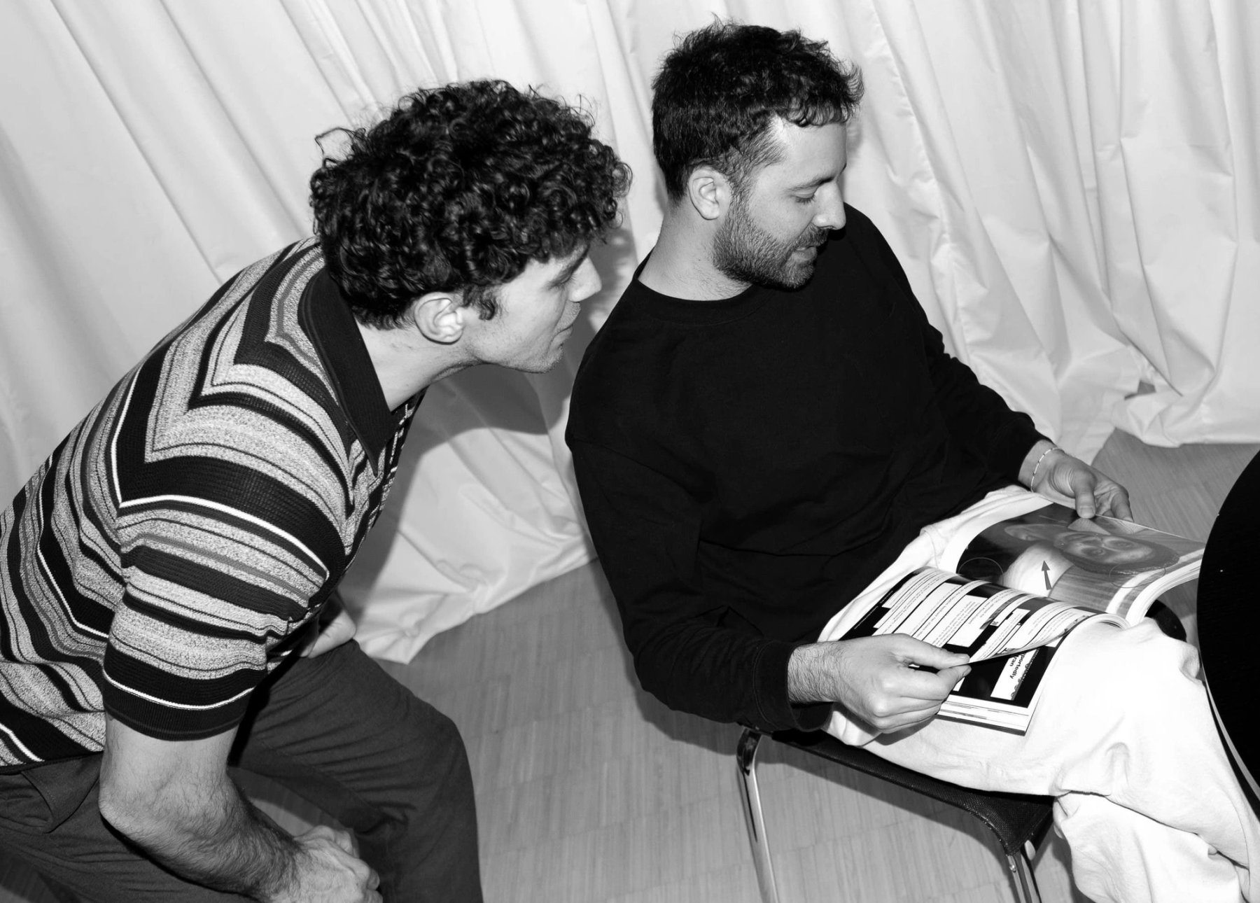 A black and white photograph of two men looking at a magazine together.