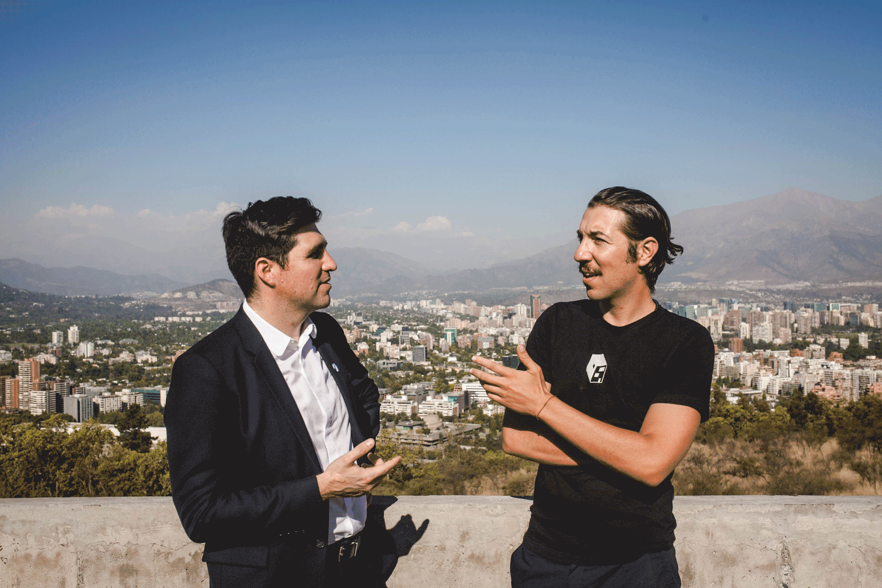 Two men talk on a terrace above a city with mountains behind it.