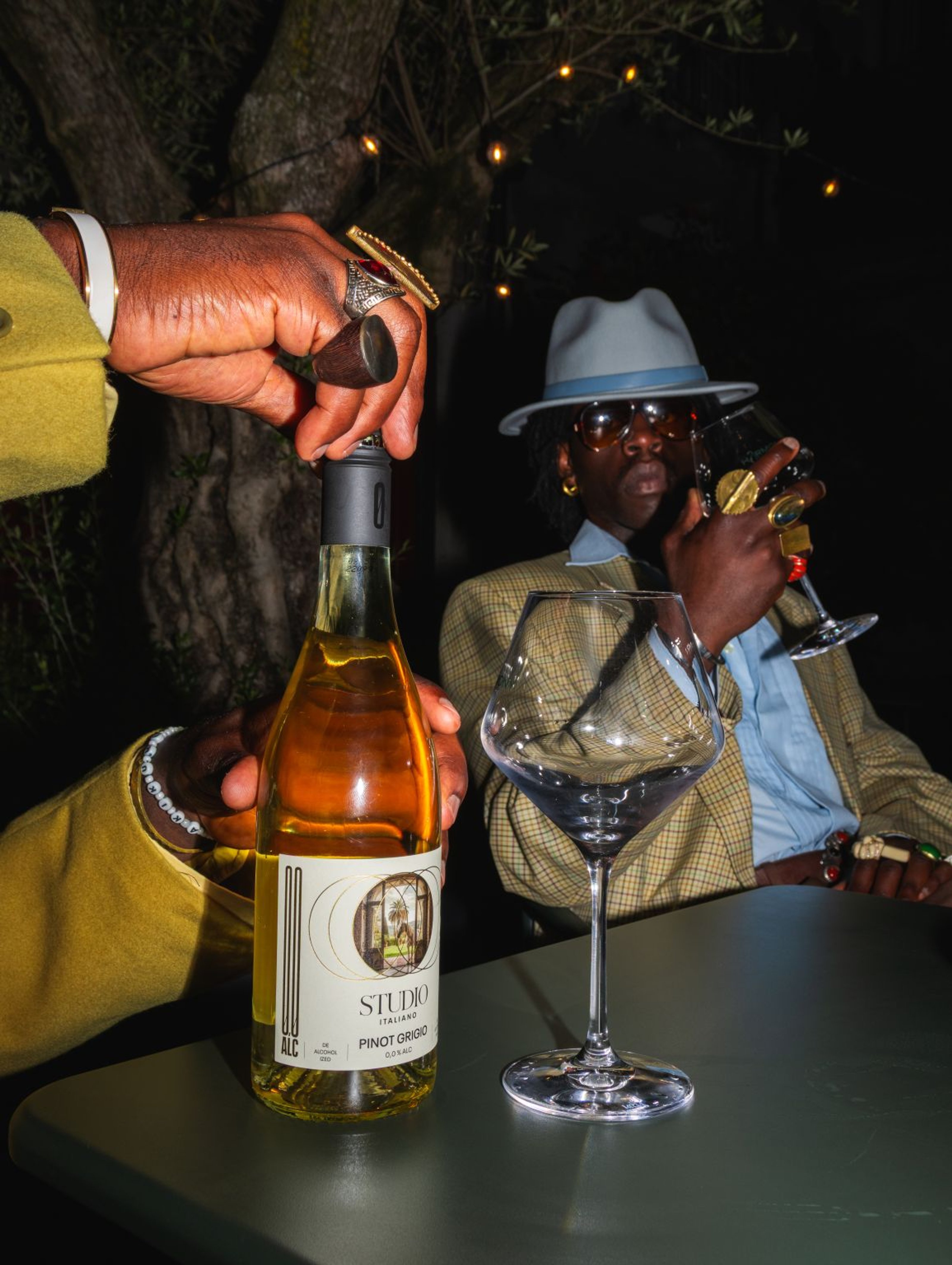 A hand pours from a bottle of Studio Italiano Pinot Grigio, a man in a hat and sunglasses behind.