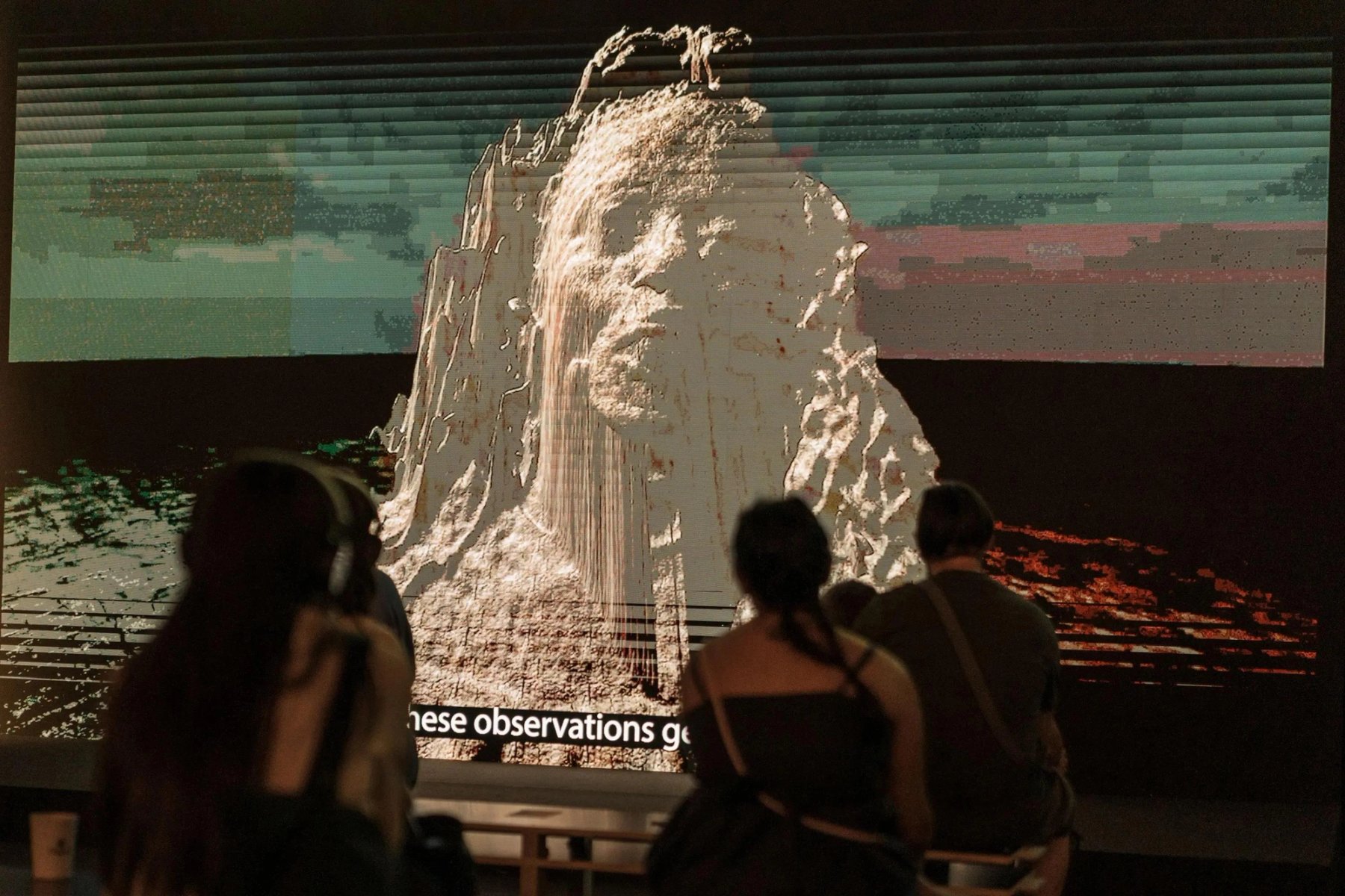 Visitors watch a projection behind a large white model of a rock formation.
