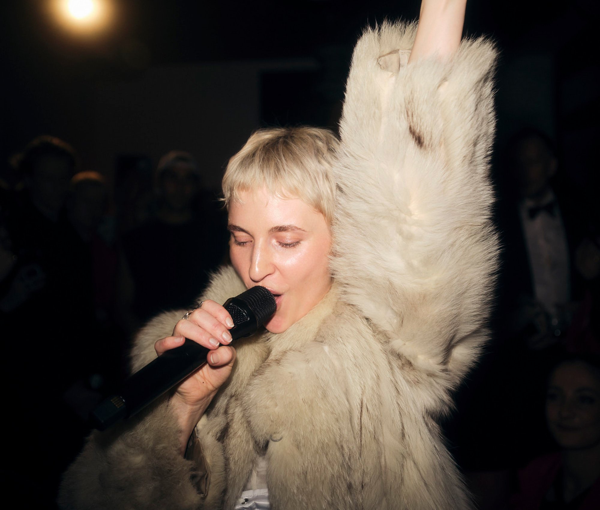 A singer in a fur coat sings into a microphone with one arm raised.