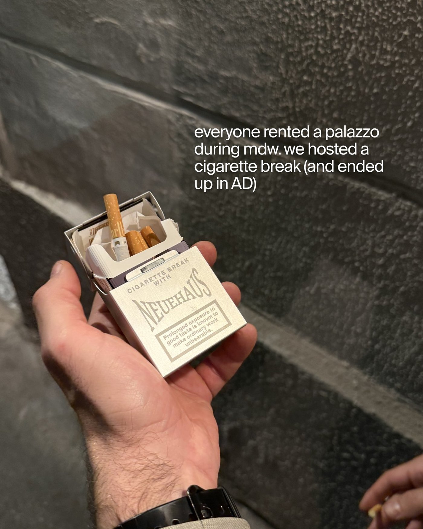 A hand holds an open NeueHaus cigarette pack against a stone wall.