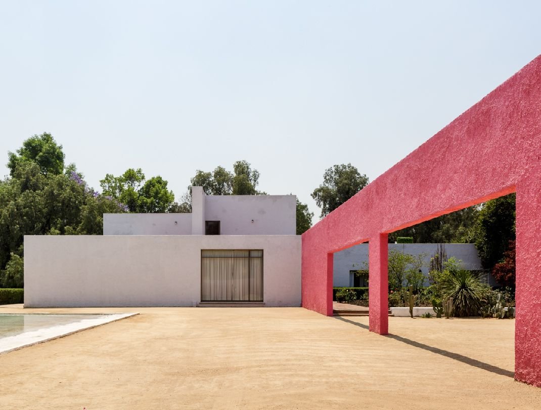 A white house behind a bright pink wall, with a pool in front of it.