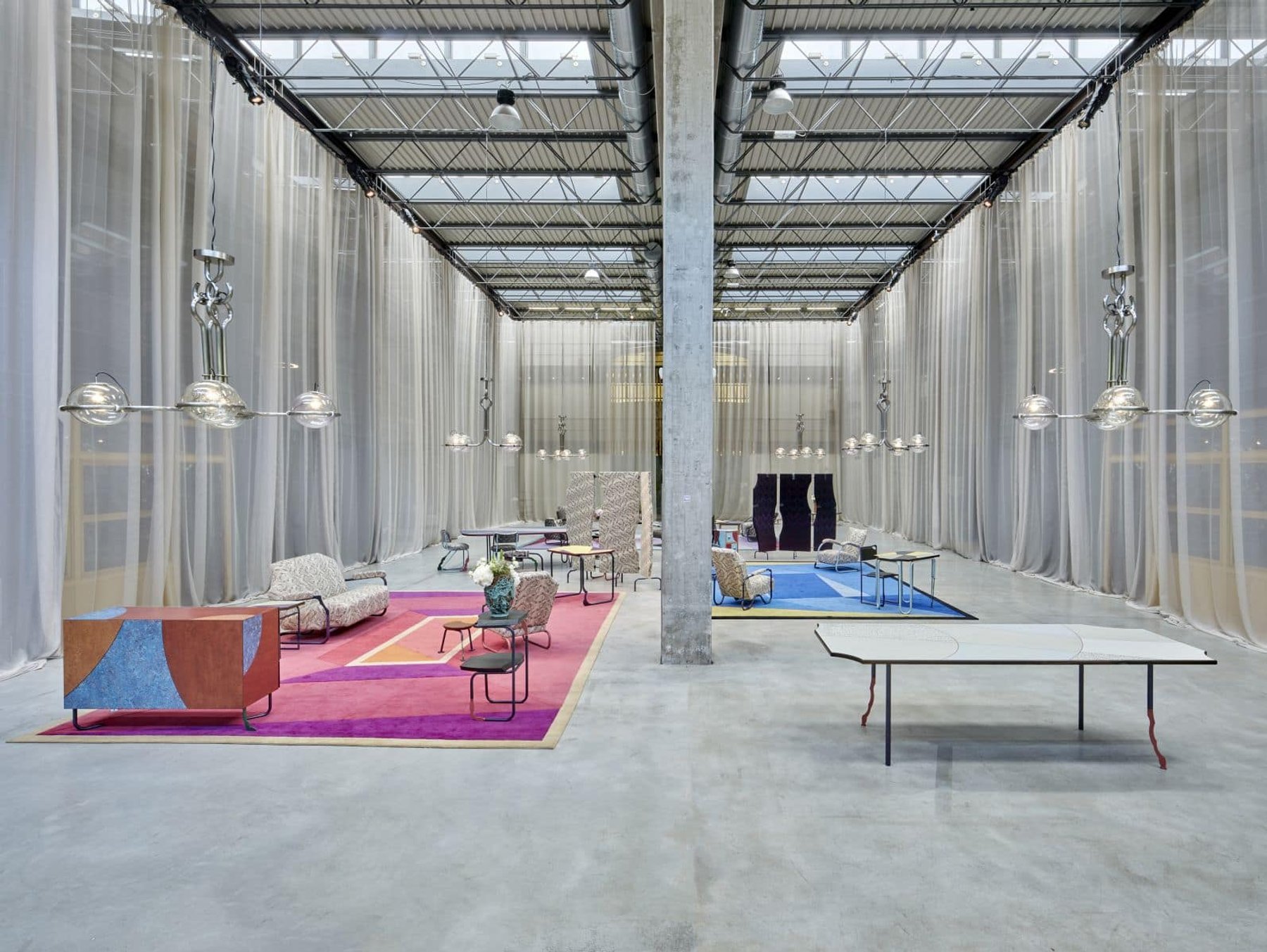 A design fair in a factory hall, with pink and blue carpets and sheer curtains between the columns.