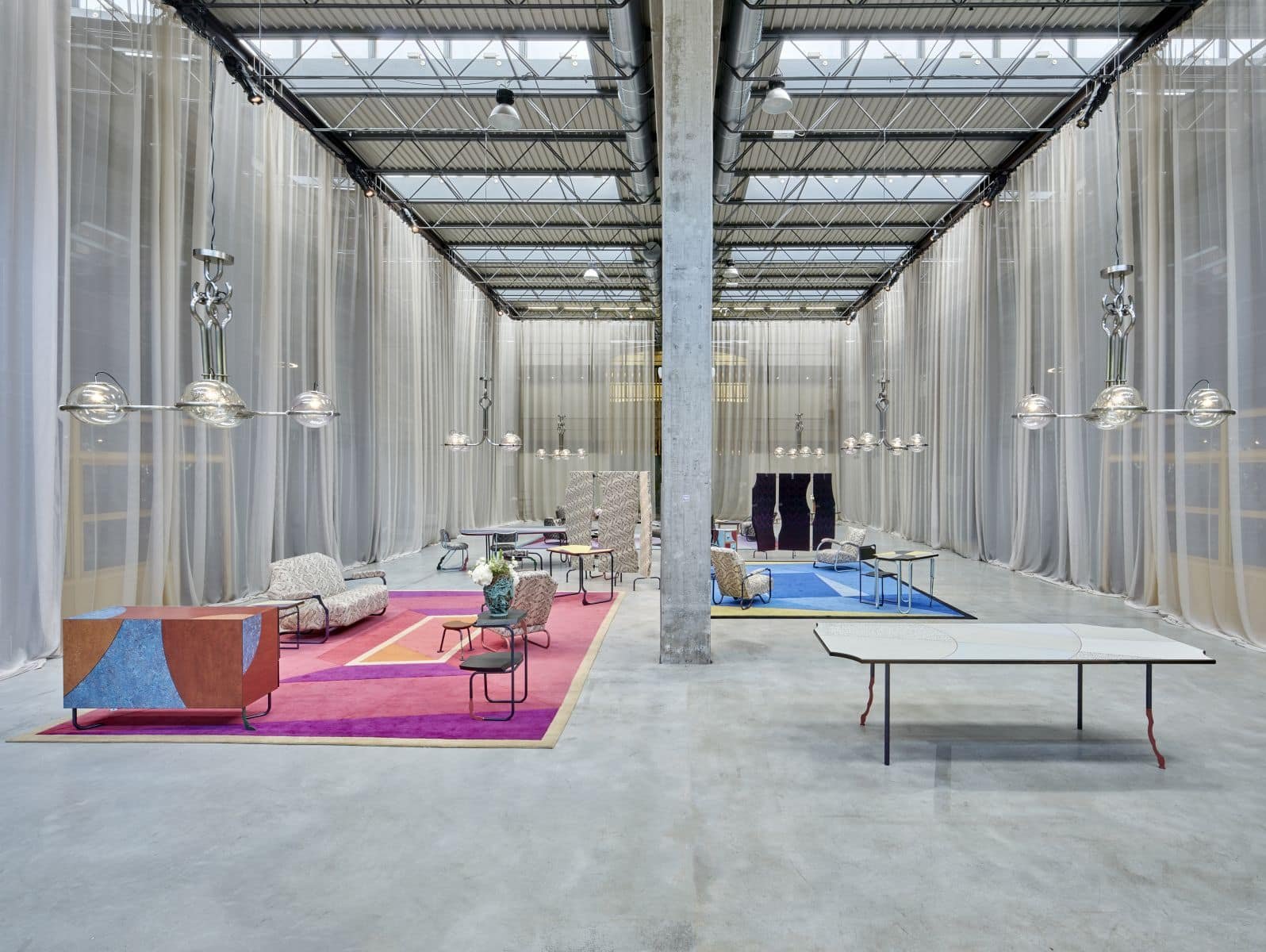 A design fair in a factory hall, with pink and blue carpets and sheer curtains between the columns.