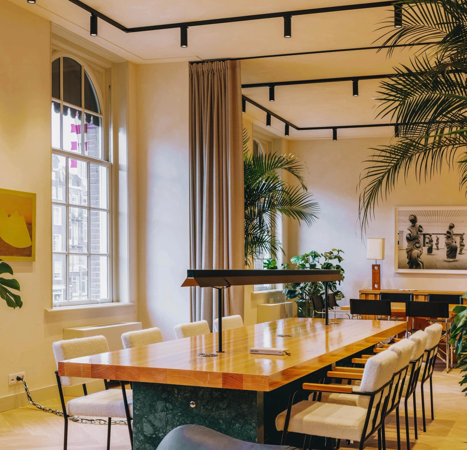 A long wooden meeting table in a bright room with tall windows and palms.