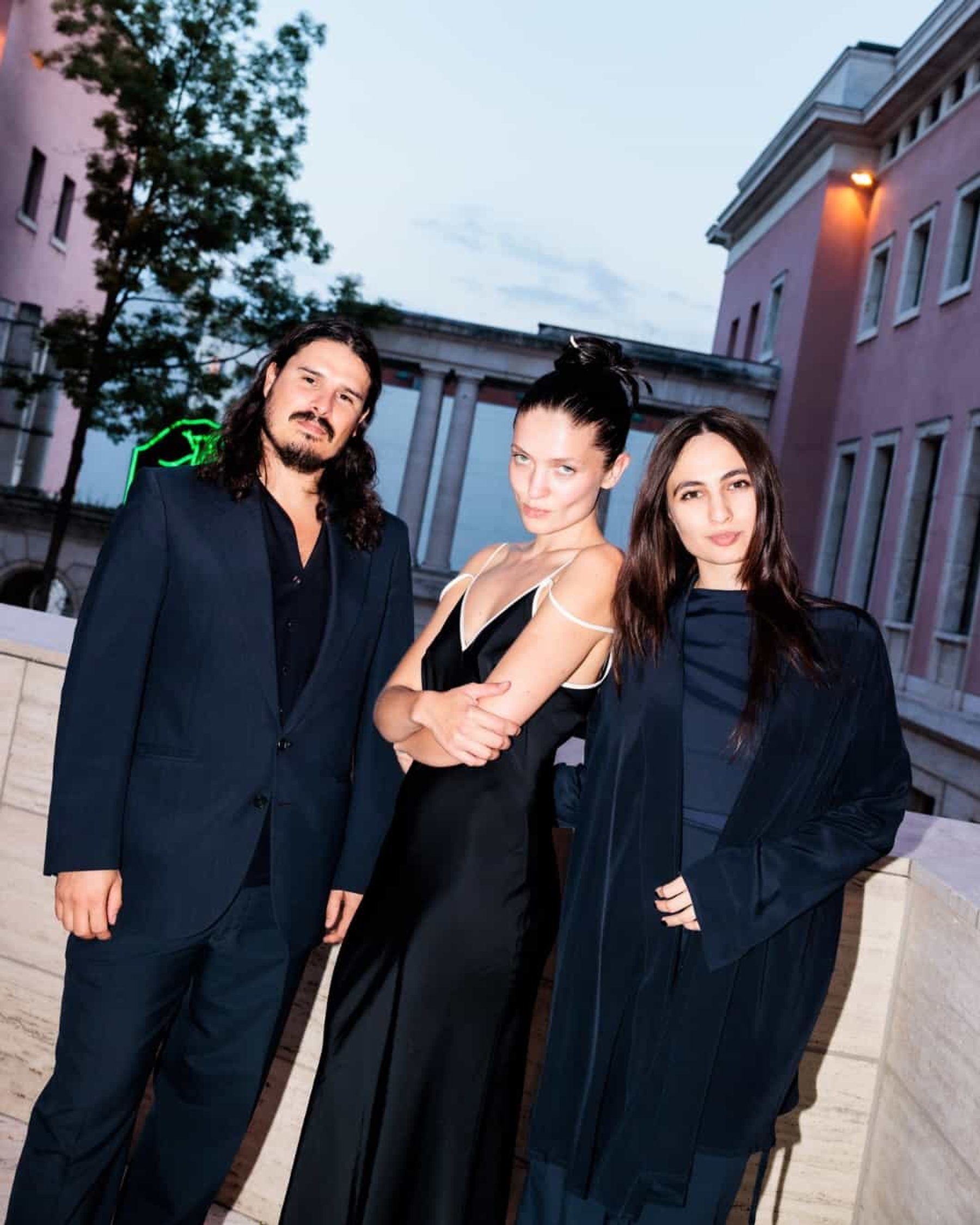 Three guests in black stand outside a pink building at dusk during art week.