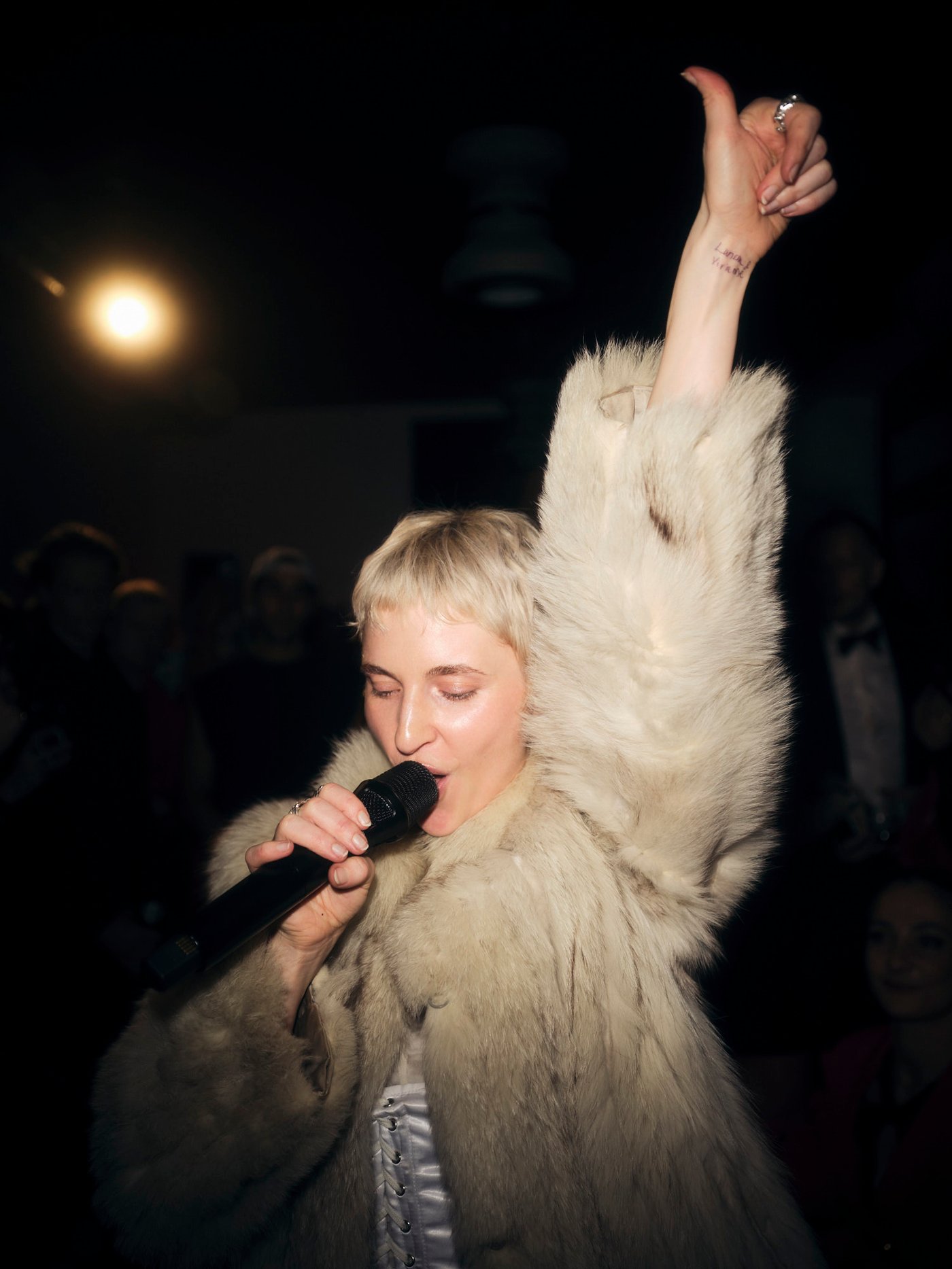 A singer in a fur coat sings into a microphone with one arm raised.