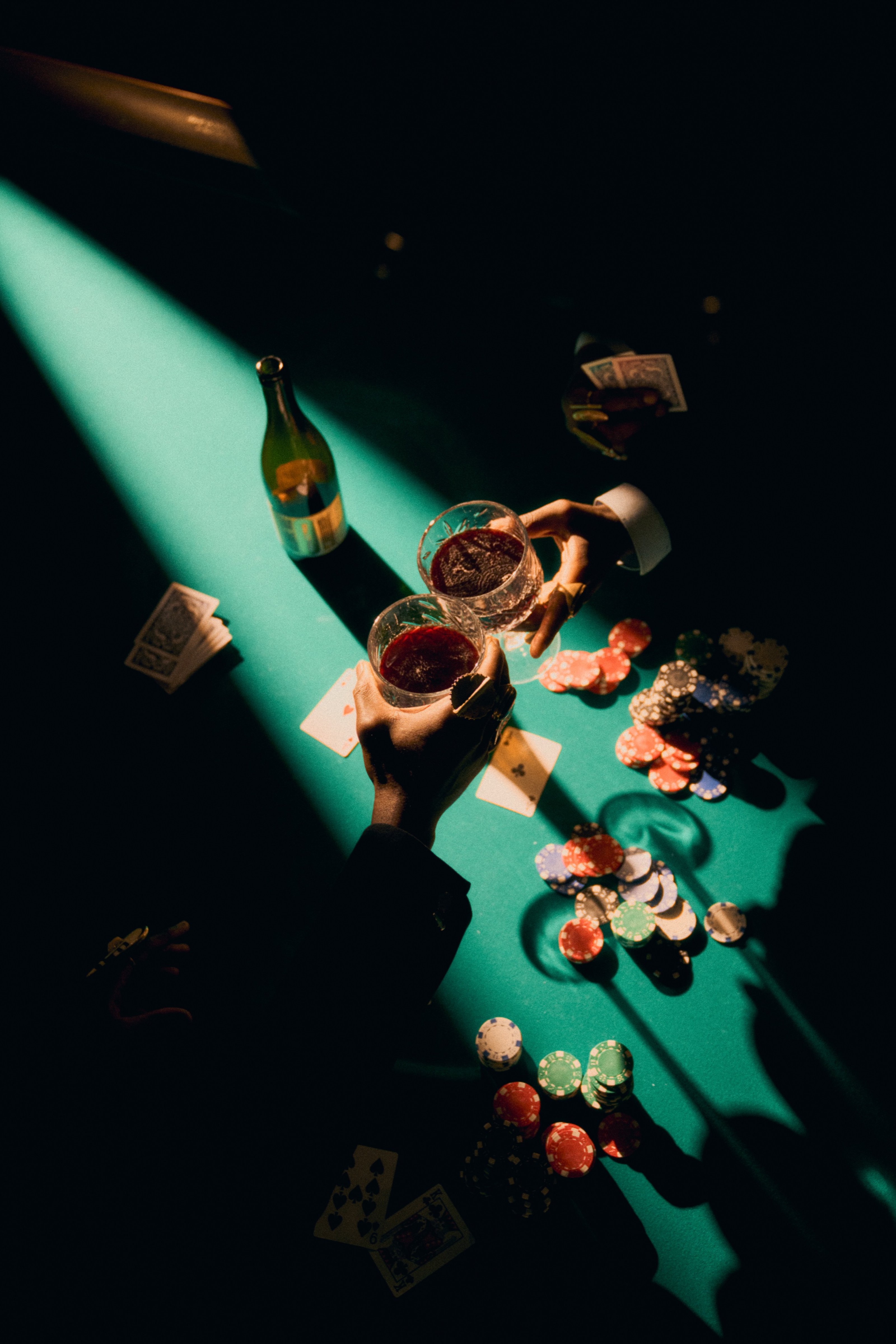 Hands reaching for glasses of red wine and poker chips in a beam of green light.