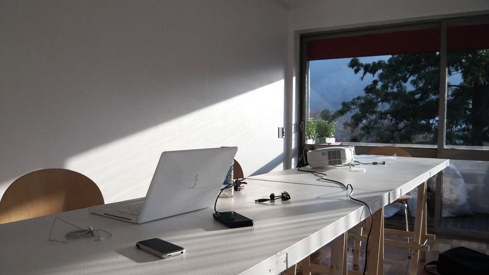 A laptop and a projector on a white desk in the sun, beside an open window.