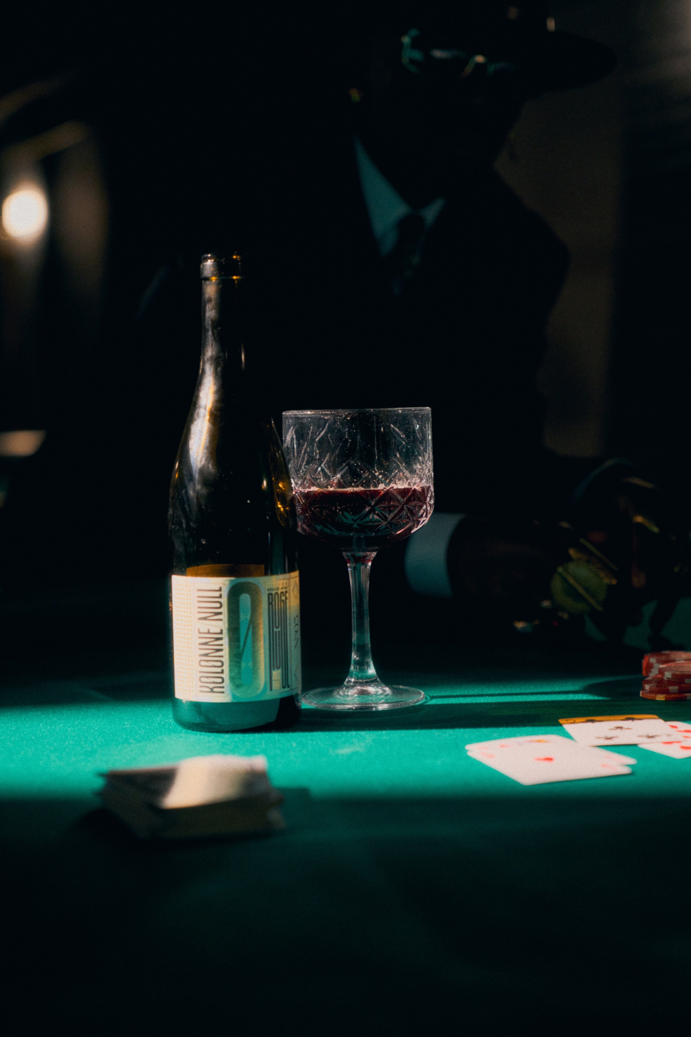 A bottle of Kolonne Null and a glass of red wine on a green card table, under a beam of light.