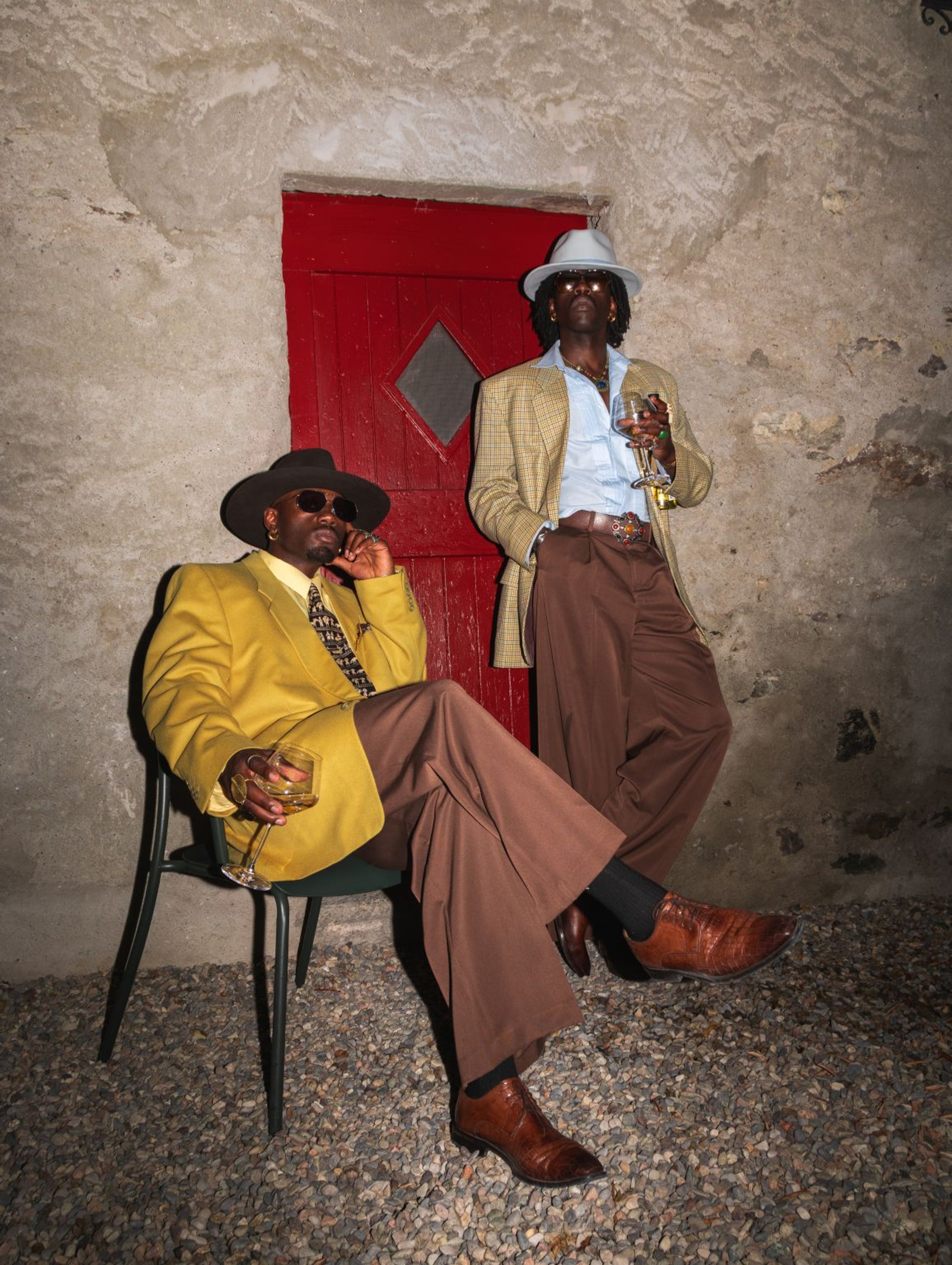 Two men in tailored suits and hats pose in front of a red door.