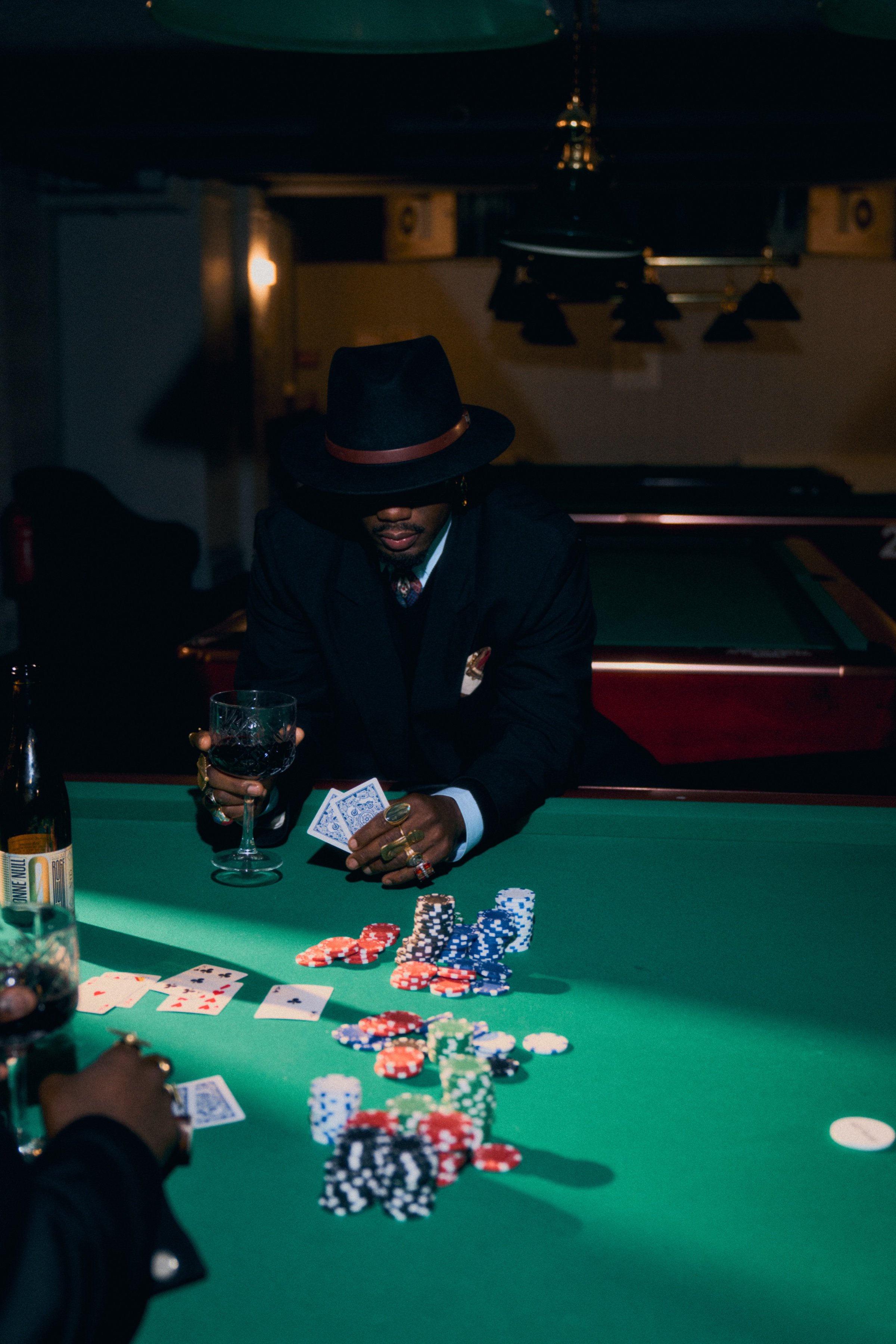 A man in a hat deals cards at a poker table, a bottle of Kolonne Null beside him.