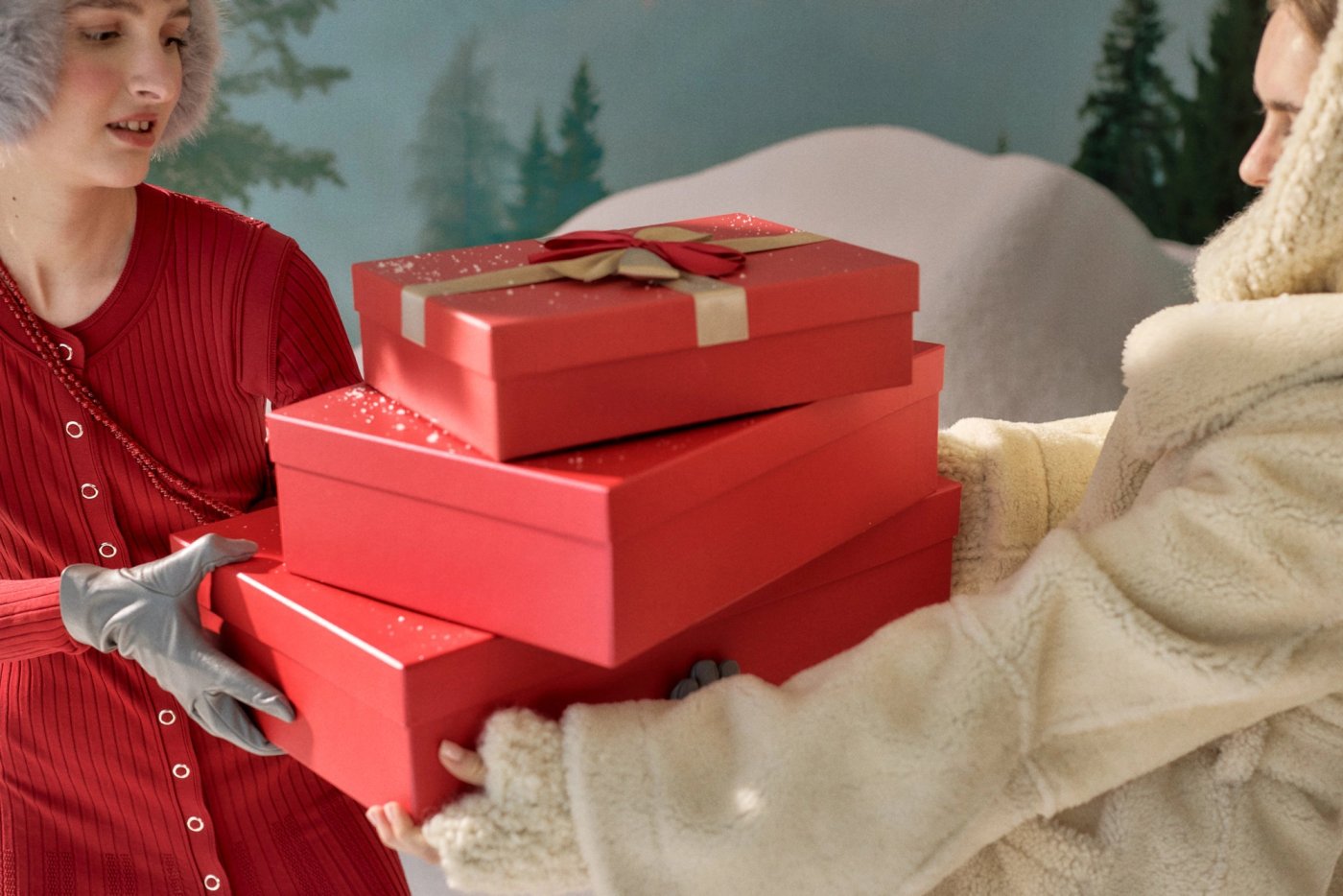 Two people in winter clothes hand over a stack of three red gift boxes tied with a ribbon, a snowy forest set behind