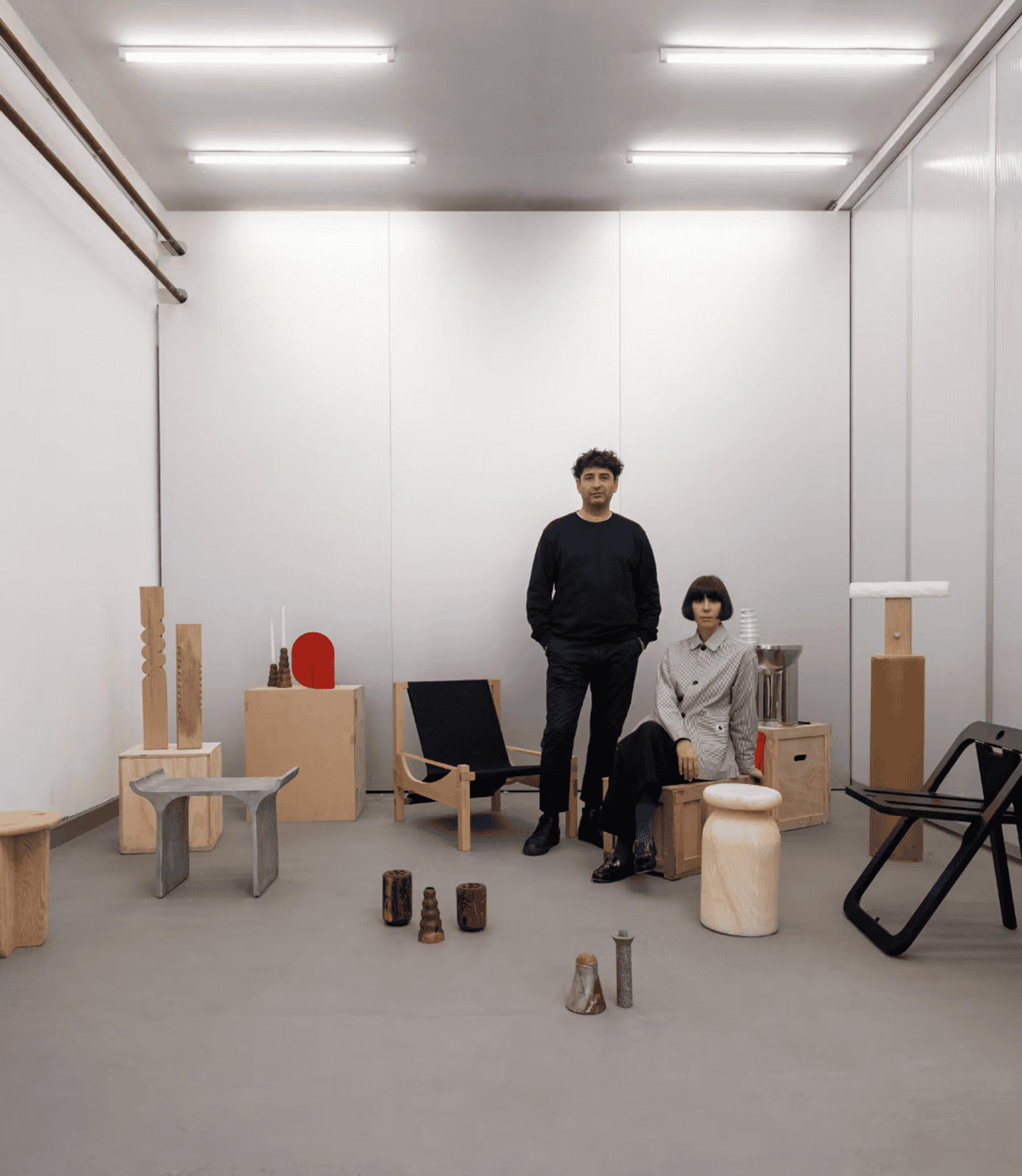 Two designers stand in a white studio among their wooden furniture pieces.