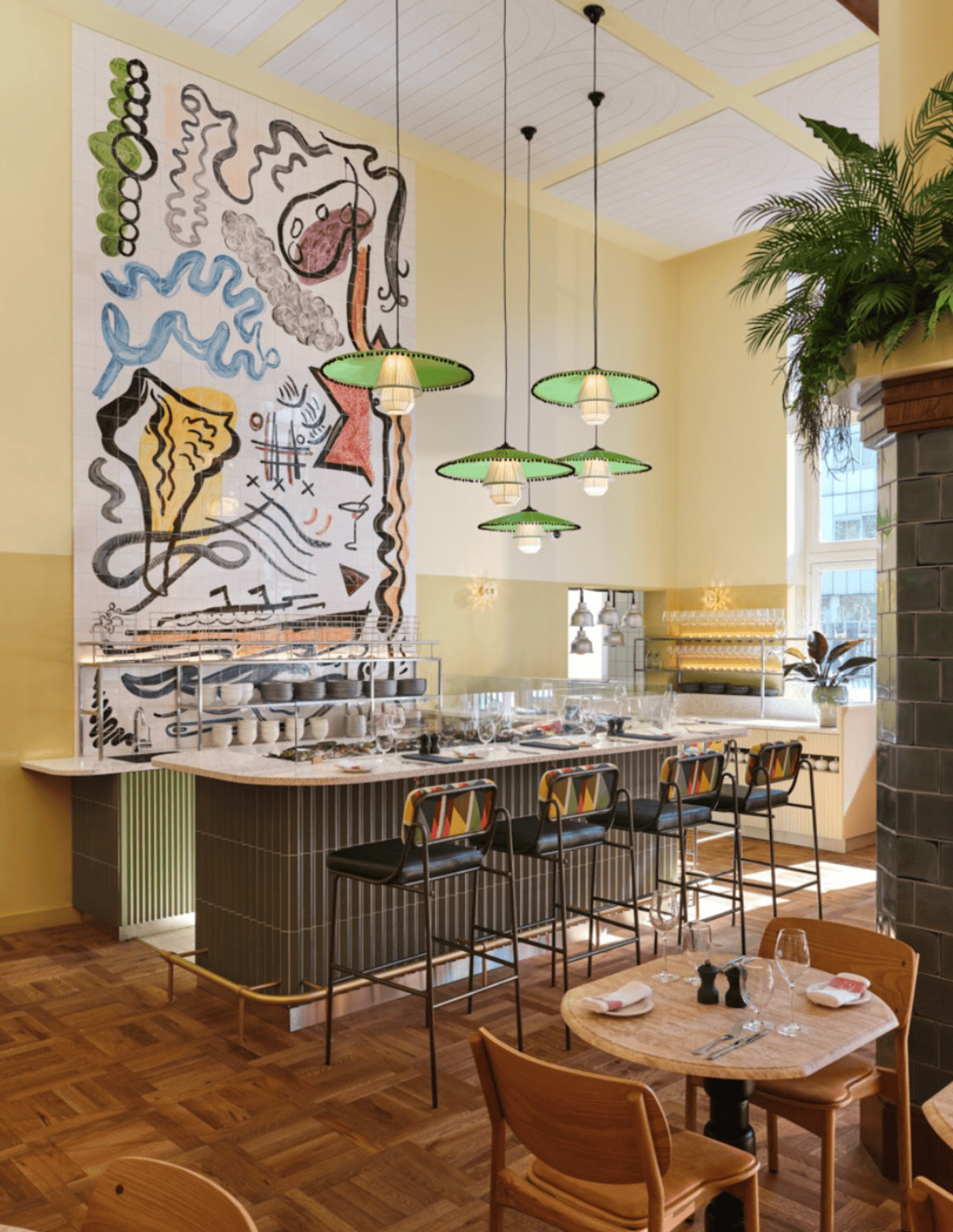 A cafe counter under a painted mural, with green glass lamps hanging over it.