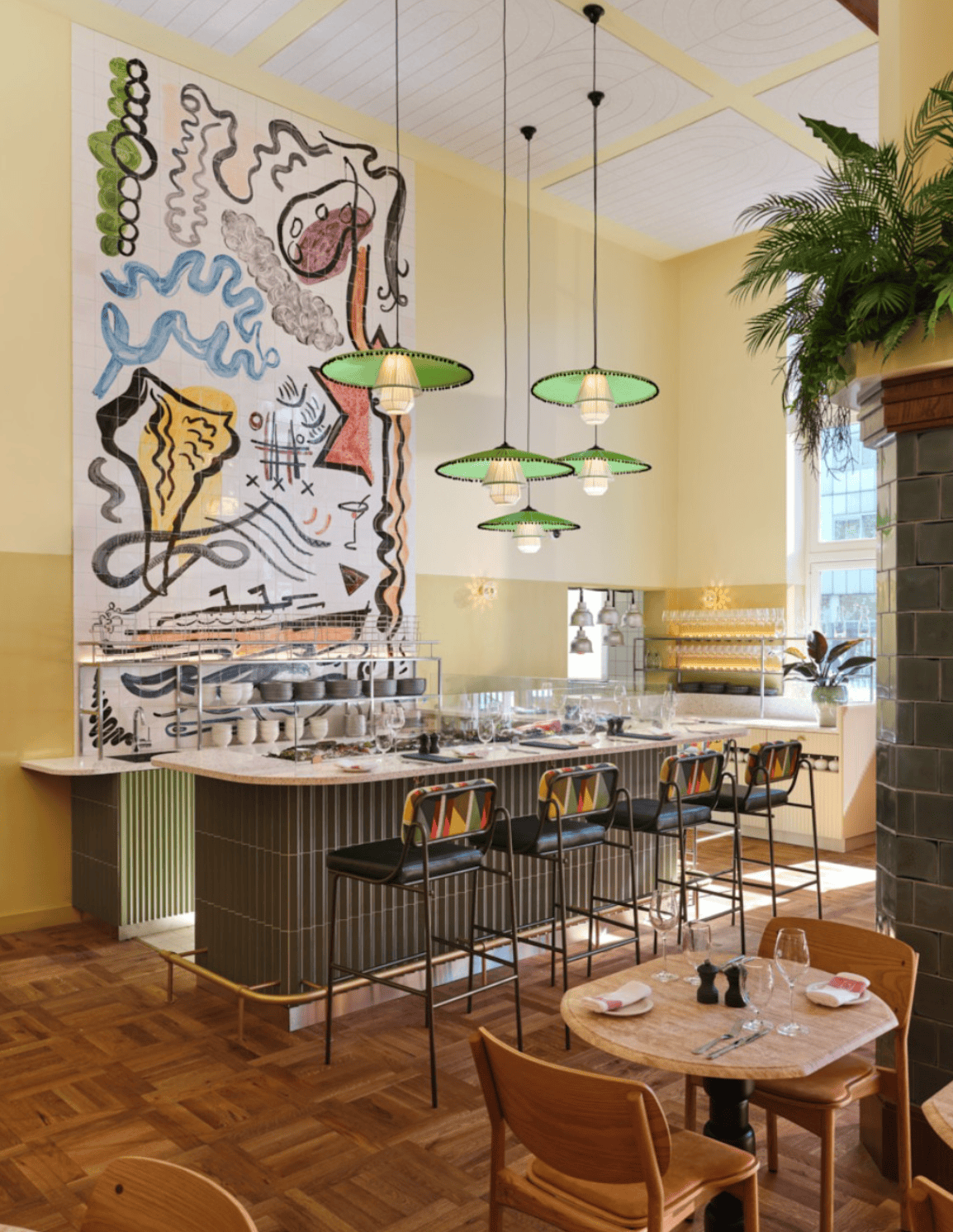 A cafe counter under a painted mural, with green glass lamps hanging over it.