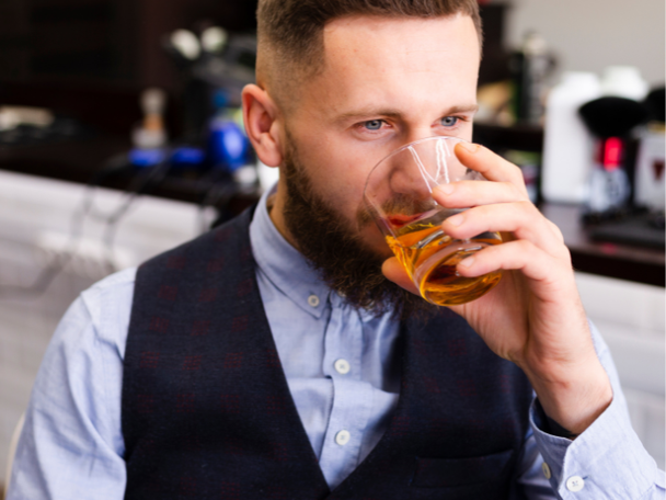 Man with beard button down shirt and vest smelling a glass of tequila.