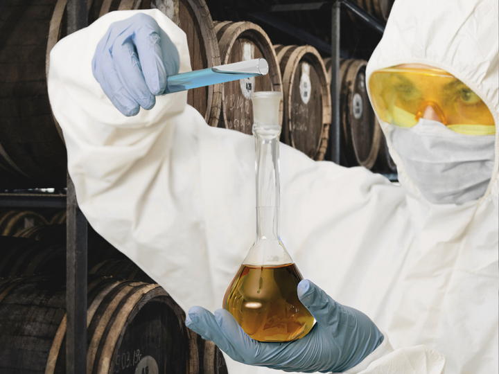 A chemist in a hazmat suit pouring a blue liquid from a test tube into a flask of tequila while standing in a barrel room