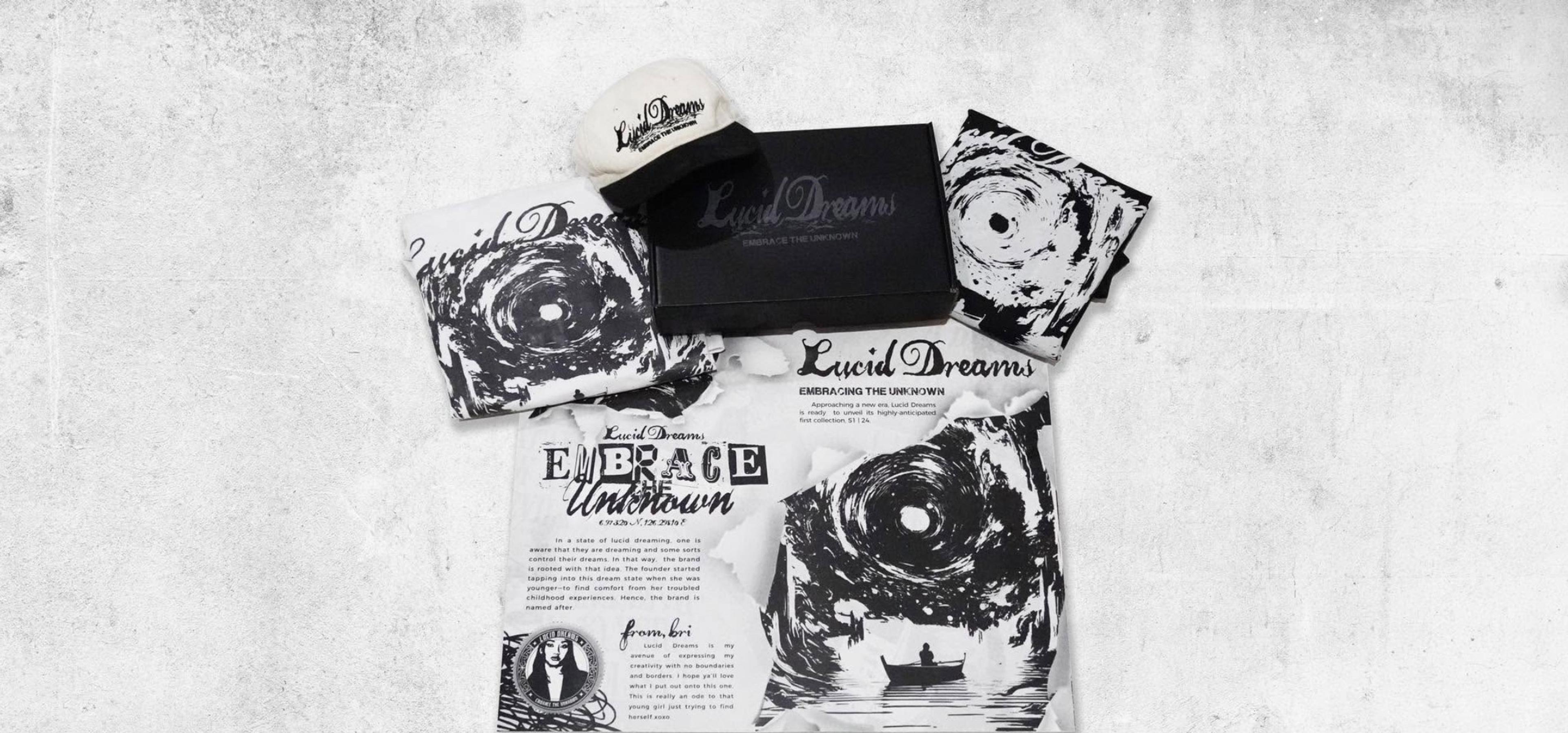 The S1 | 24 collection laid out on concrete: vortex-print tees, a two-tone cap, the black mailer box and the Embrace the Unknown zine with Bri’s note