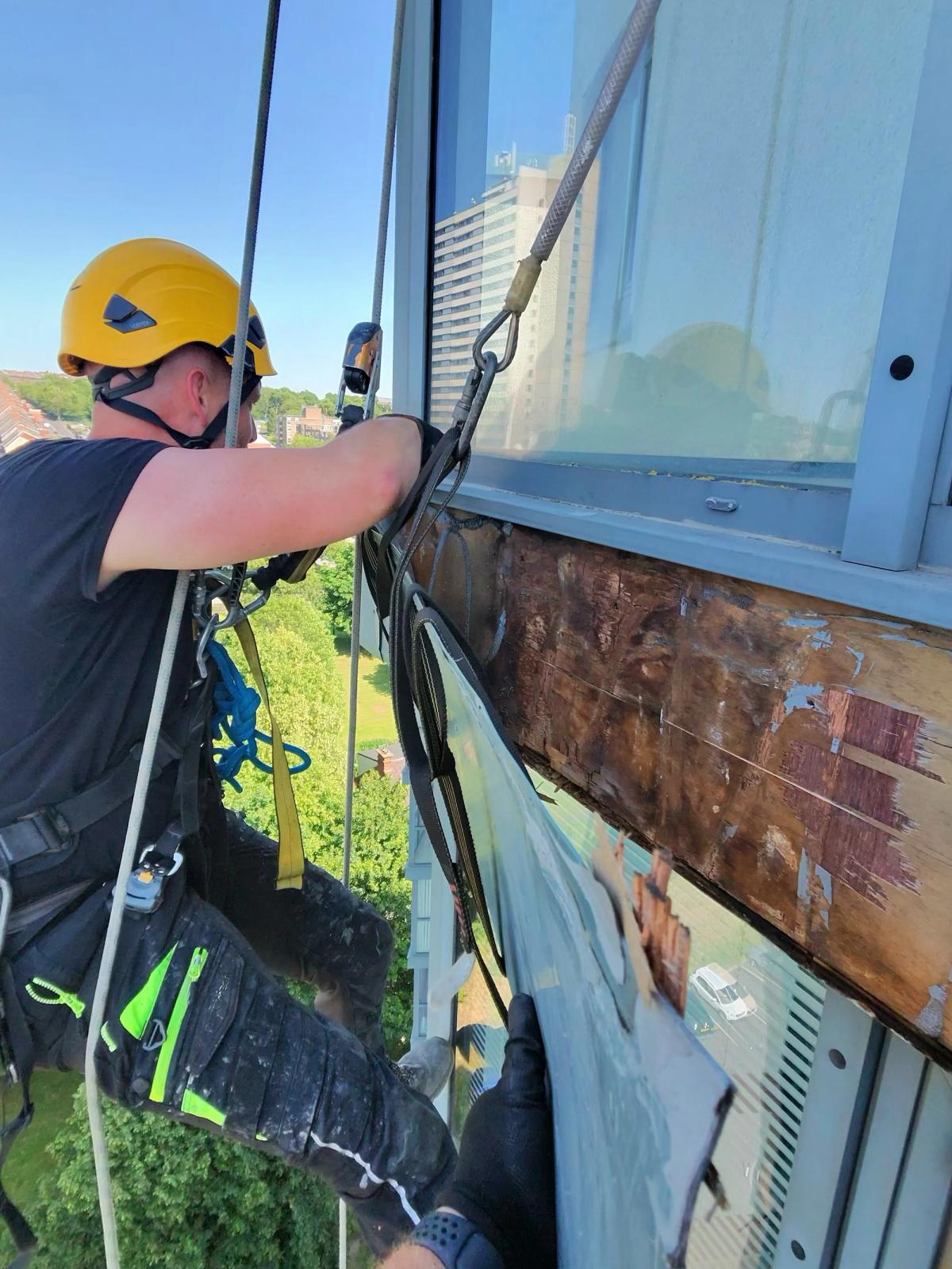 Asset Access UK rope access technician removing aluminium façade cladding from a high-rise residential building in Newcastle, exposing extensive water-damaged timber beneath the panels.