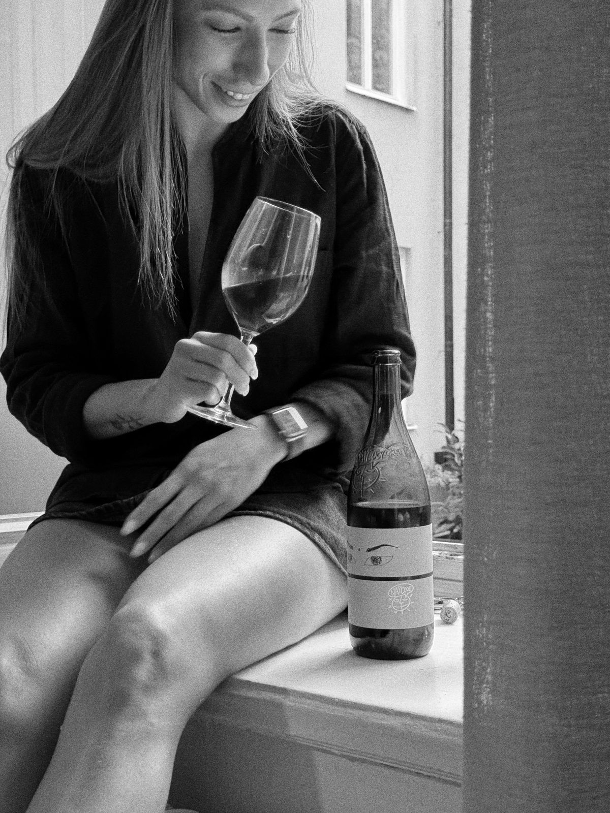 Black-and-white photo of a woman sitting by a window with a glass and a bottle of wine