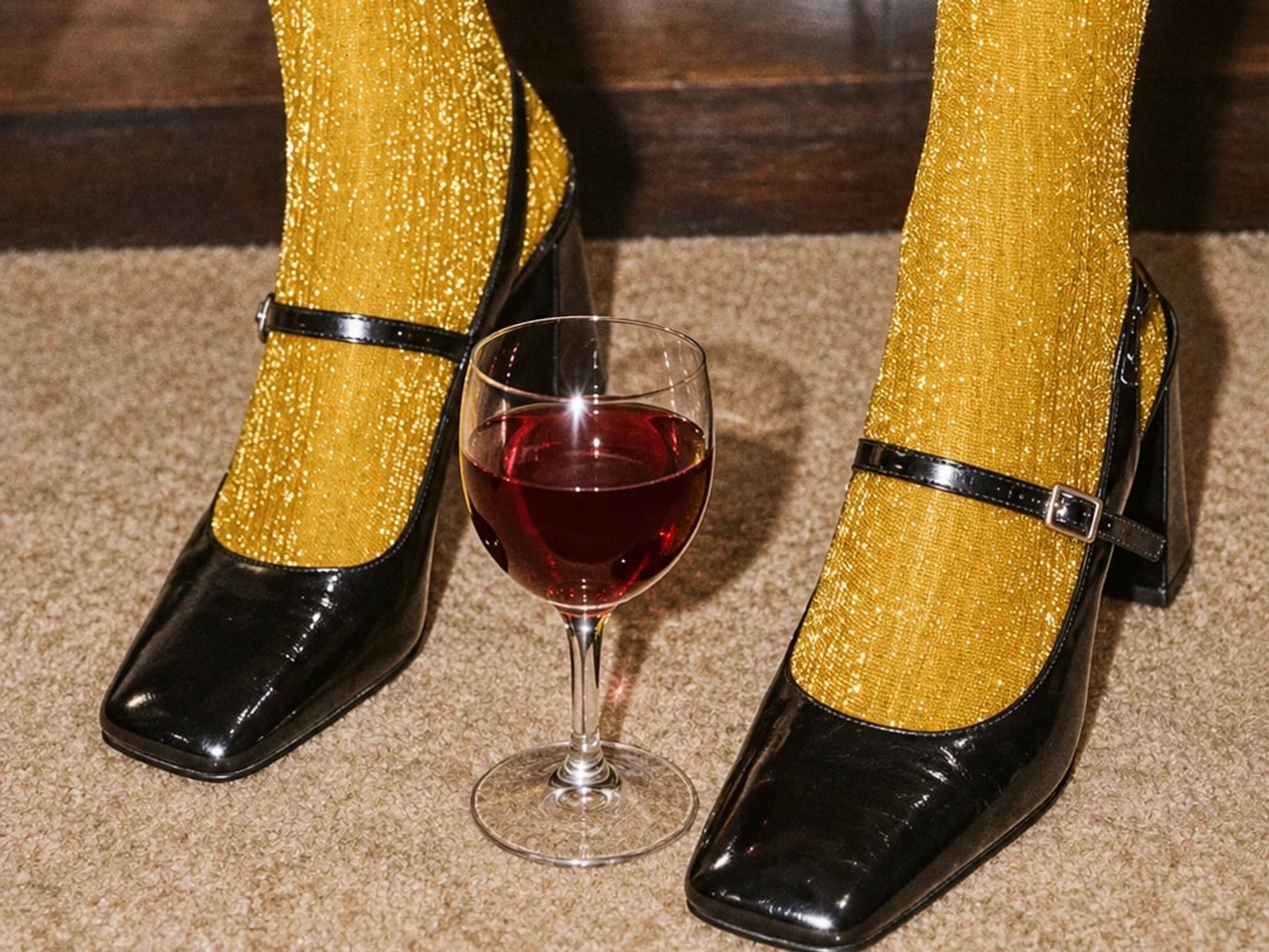 A glass of red wine on a carpet between feet in black patent Mary Jane shoes and gold glitter tights