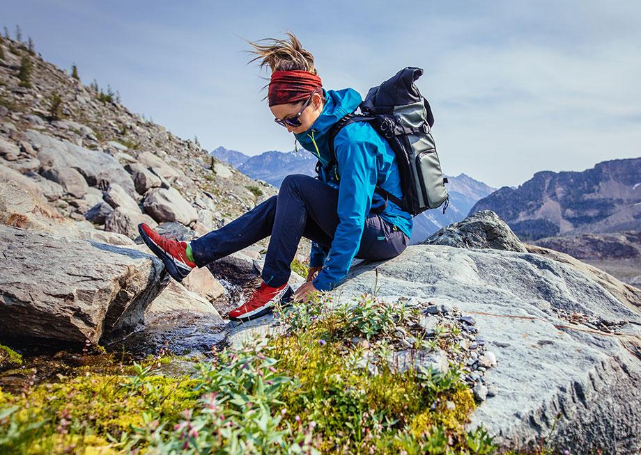Female hiker sitting on a rocky surface with a backpack, adjusting her shoe in a mountainous landscape.