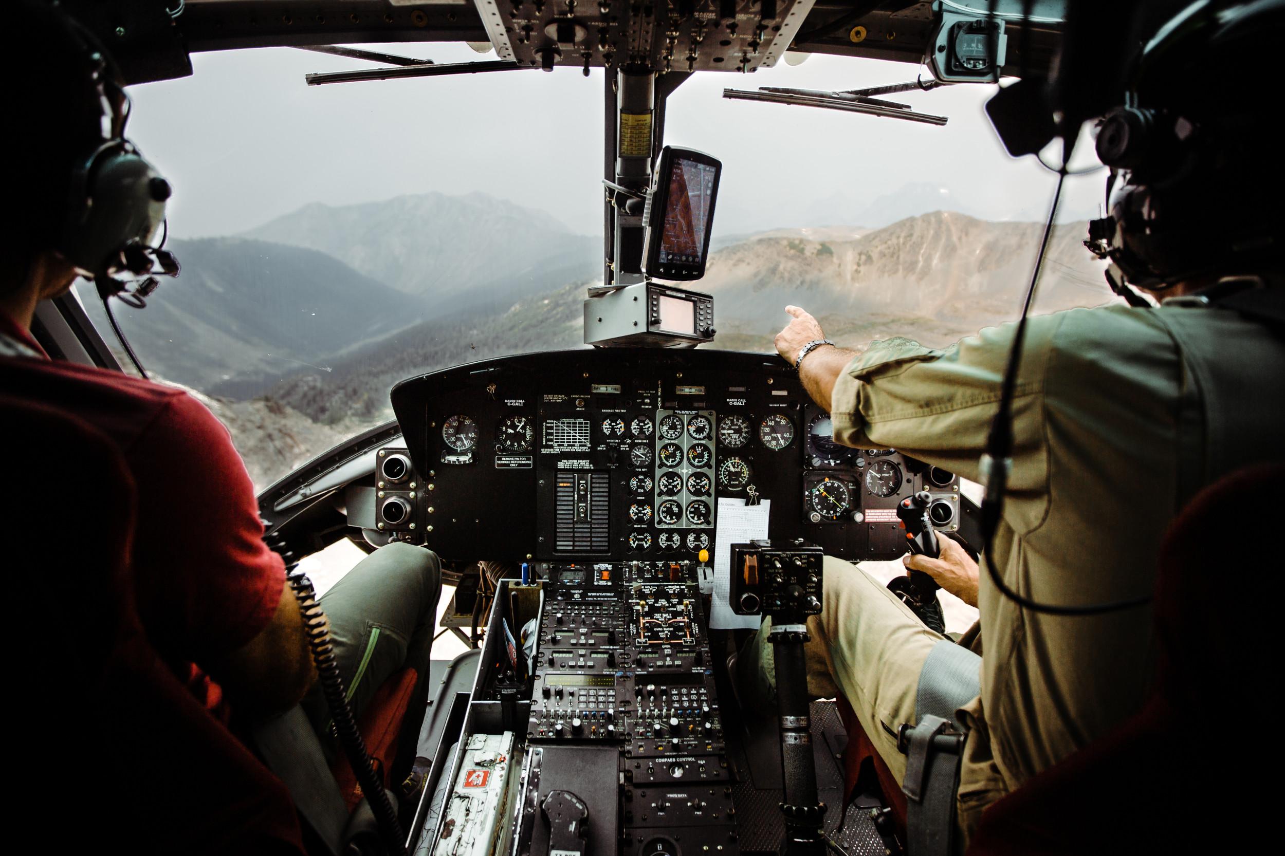 Looking into the cockpit of a helicopter, pilot pointing out the windshield at the surrounding mountains